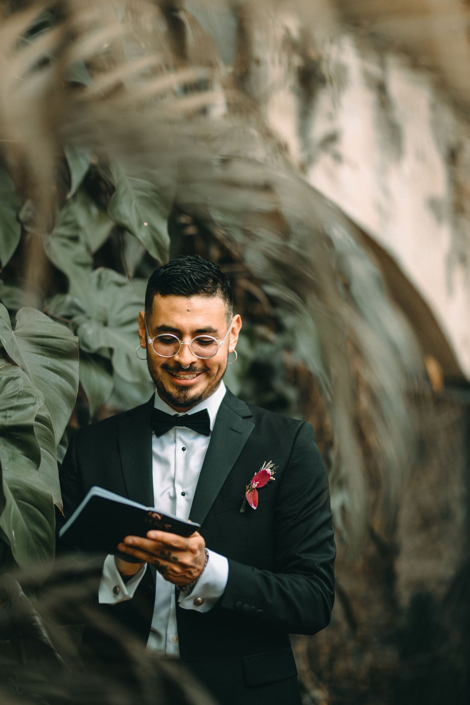 Well-dressed person in a black tuxedo and bowtie smiles while reading from a book among tropical greenery.