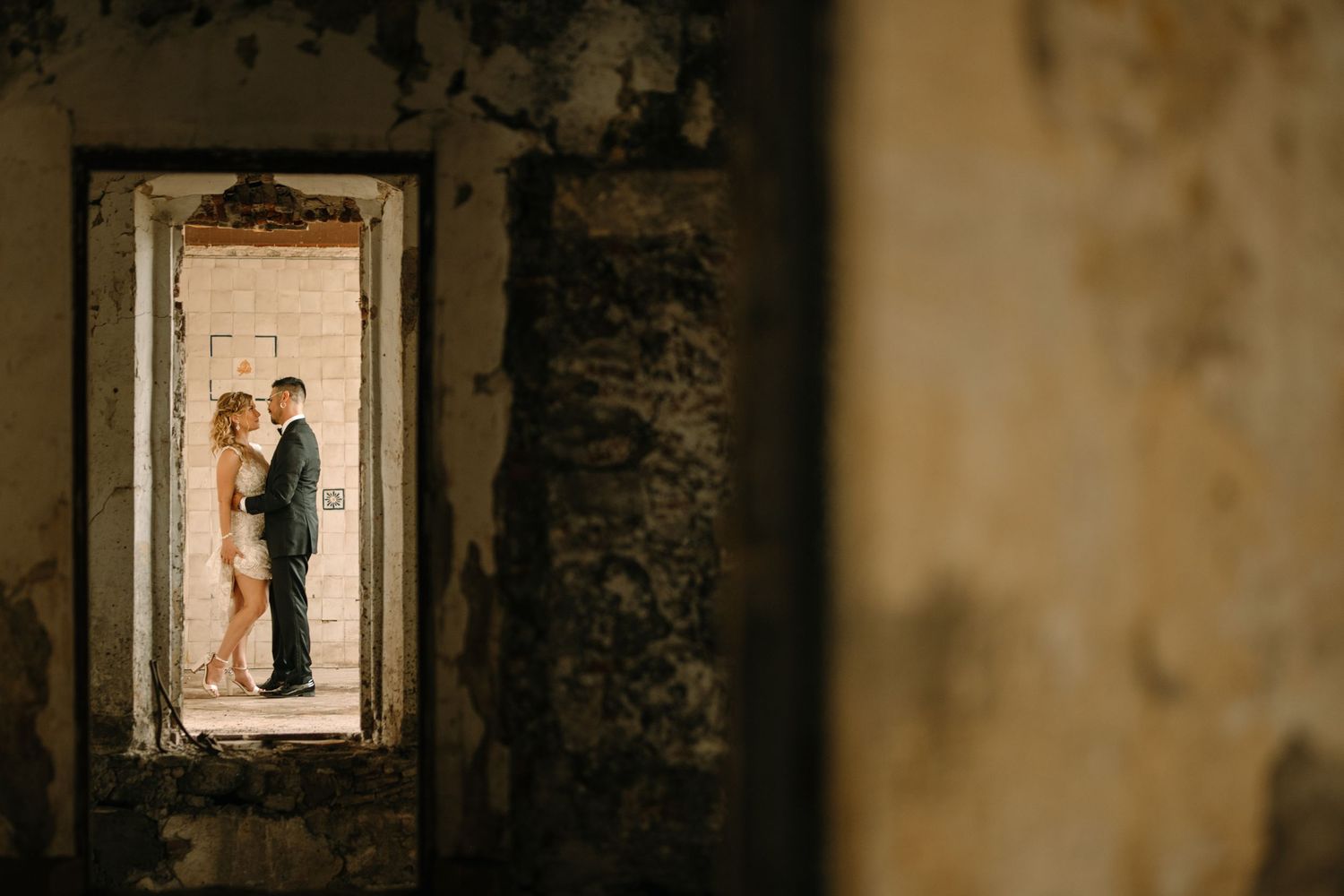 A romantic couple stands silhouetted in a weathered doorframe of an old building, creating a moody atmospheric scene.