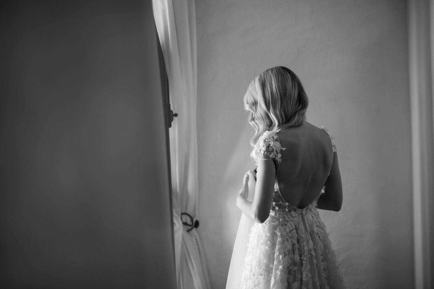 A bride in a delicate lace wedding dress stands by a window in a thoughtful black and white moment.