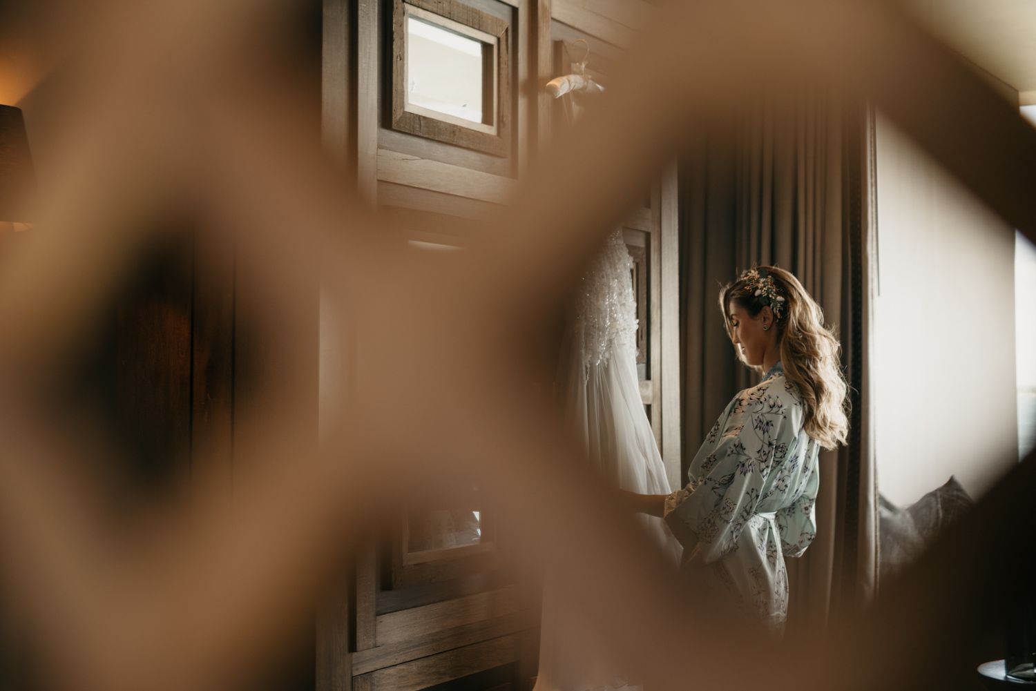A person stands in contemplative pose by a sunlit window, viewed through a blurred diamond-shaped foreground element.