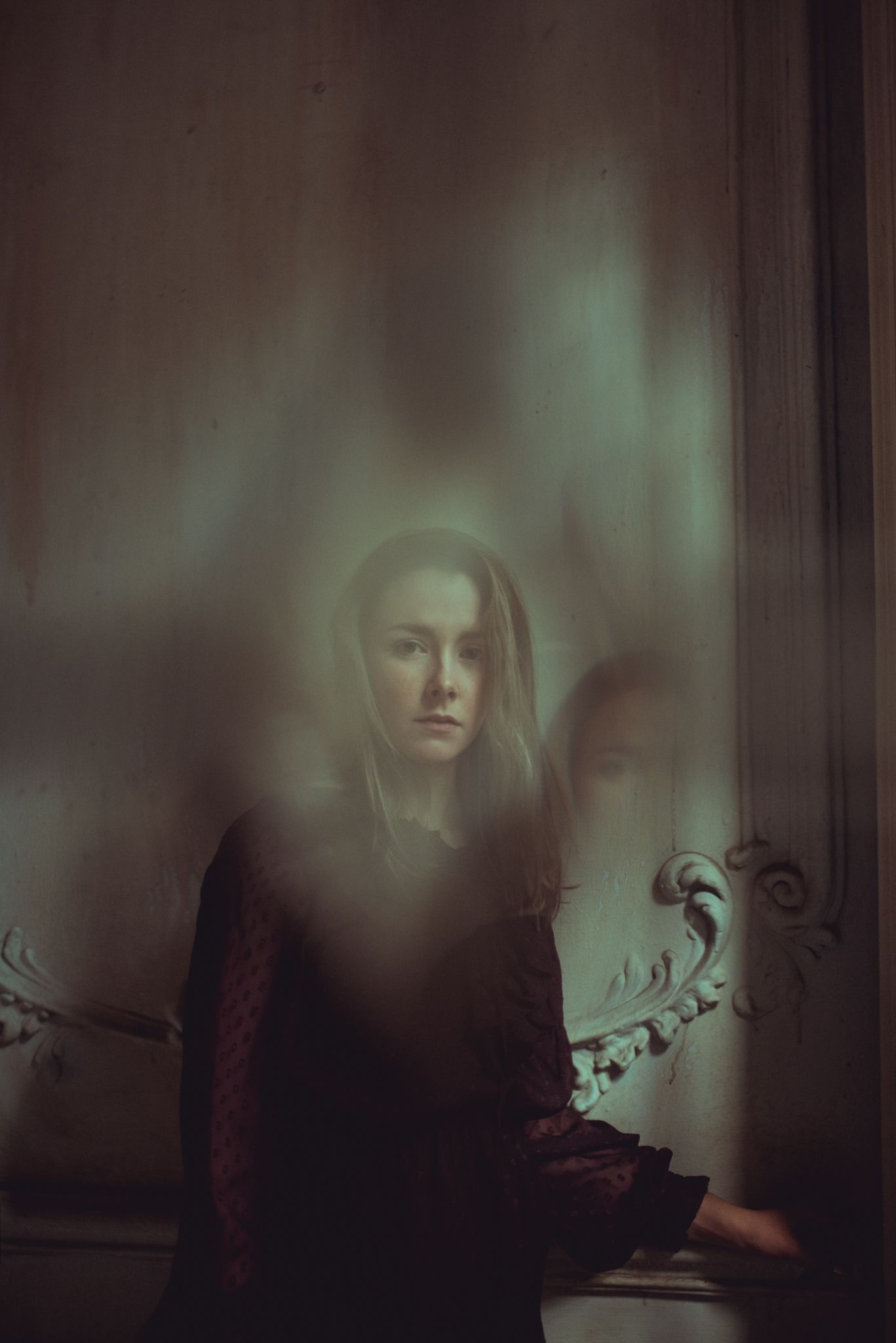 Atmospheric dark portrait with ethereal glow effect showing figure in black clothing against moody vintage background.