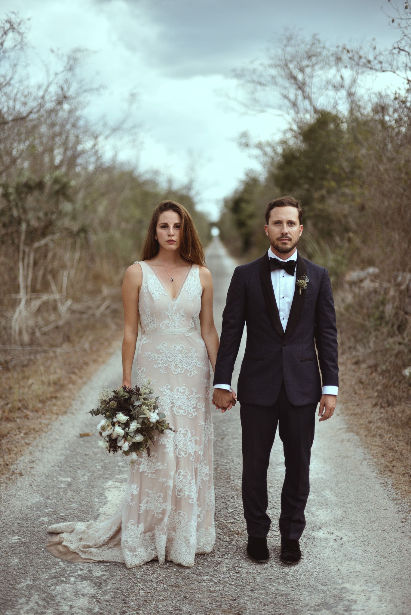A newlywed couple stands together on a country path, the bride in a lace gown holding a bouquet and groom in dark suit.