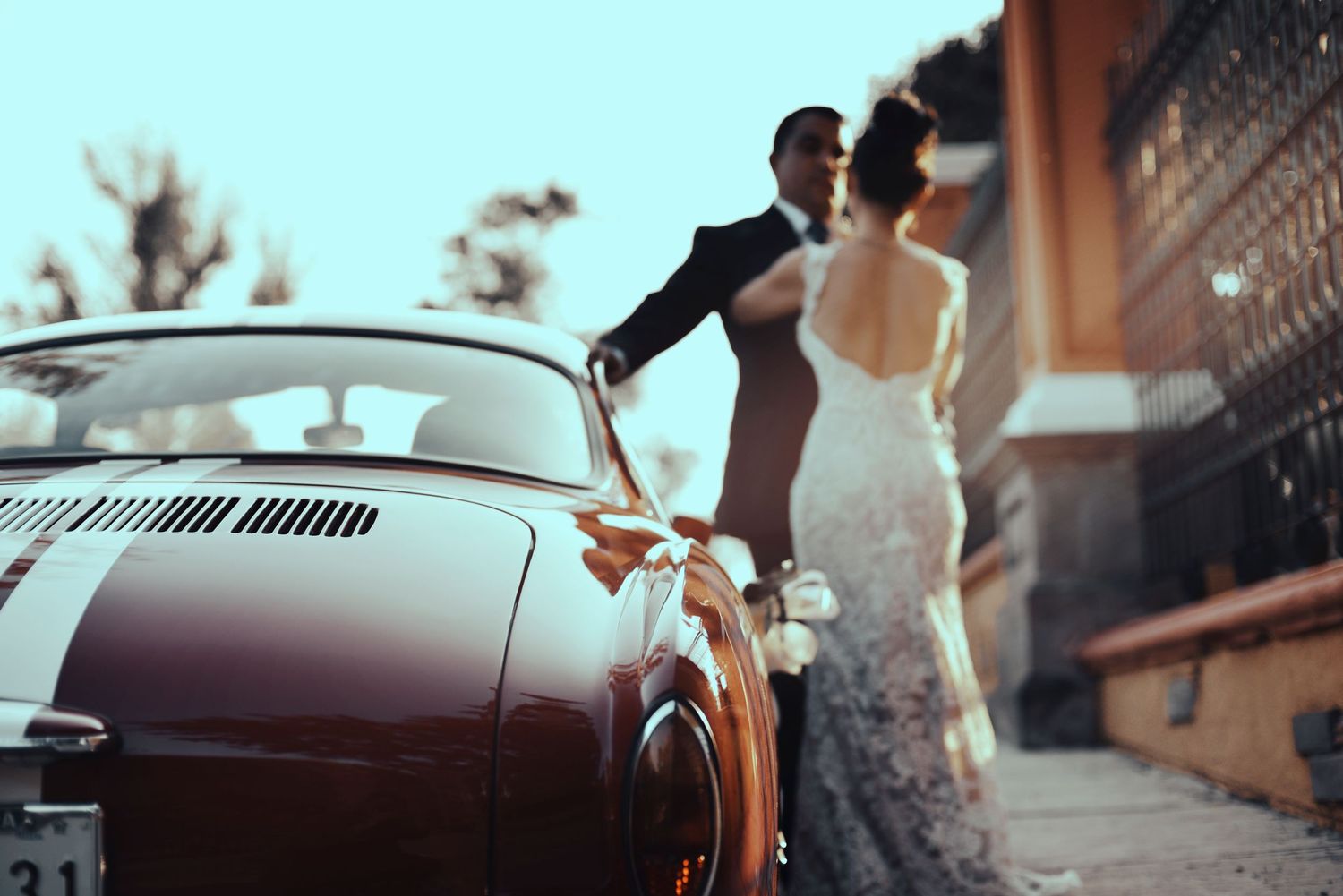 A couple stands near a classic vintage car in an elegant wedding scene with soft sunlight filtering through the air.