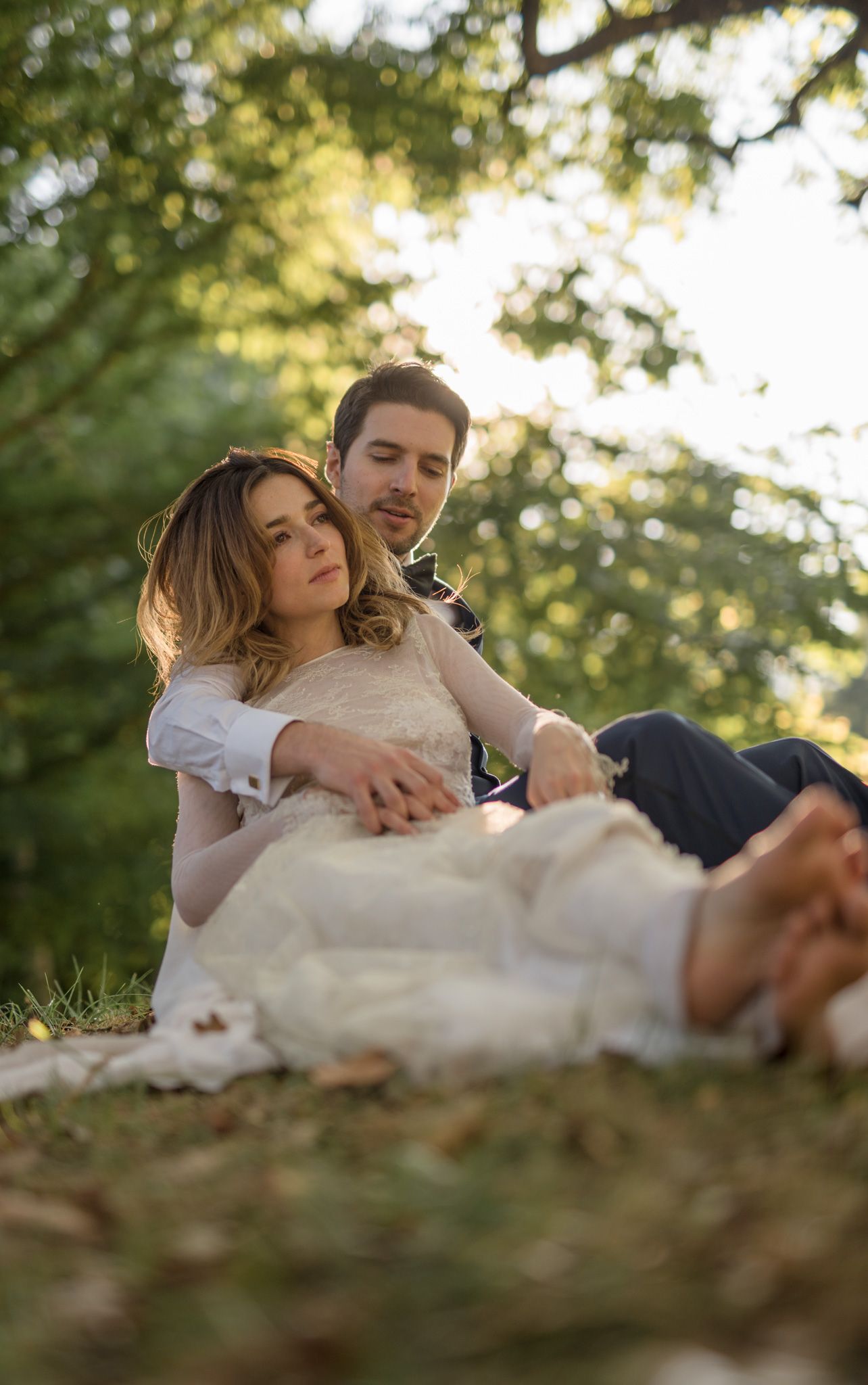 Couple relaxes together on picnic blanket in dreamy outdoor setting with sunlit trees in background.