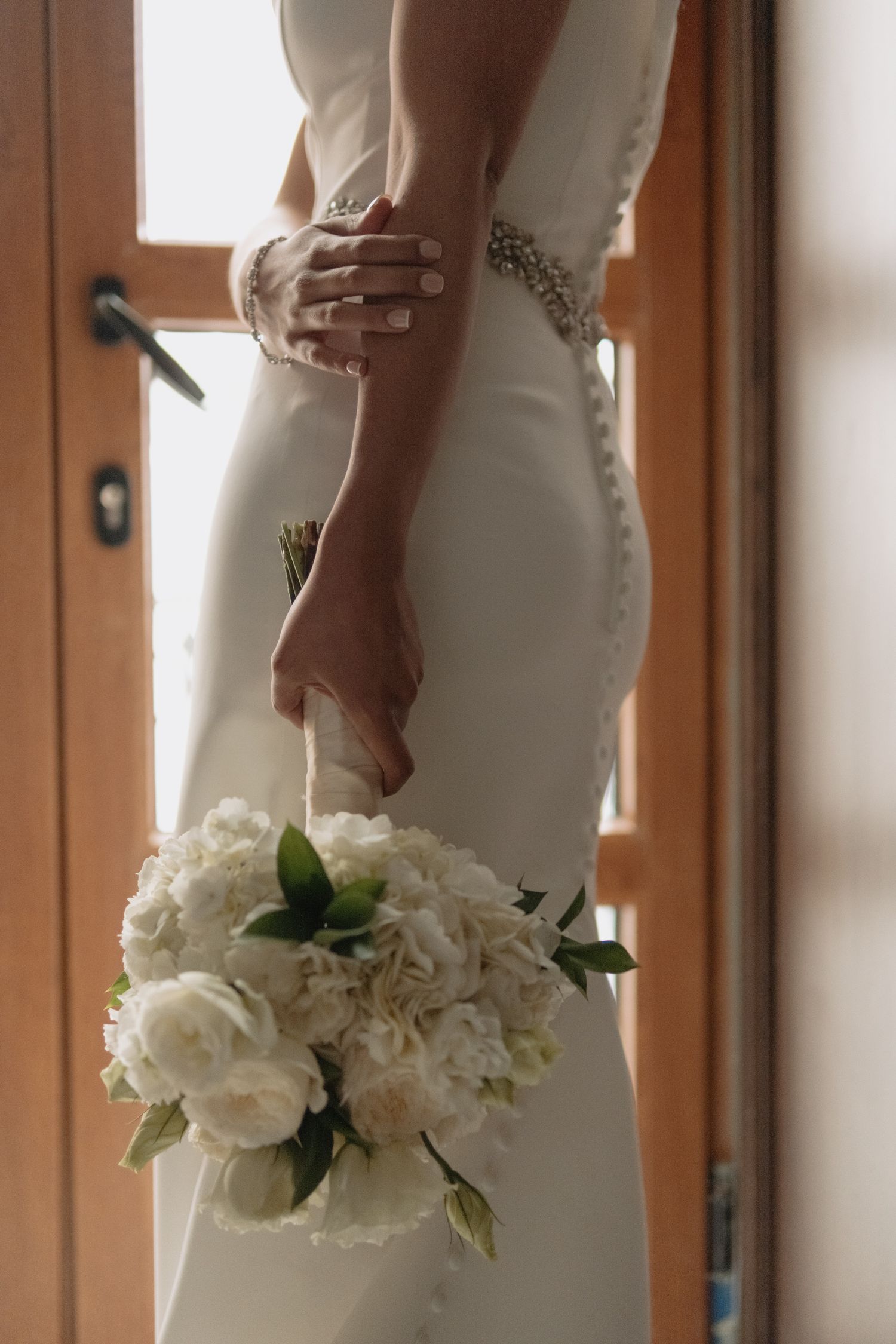 Close-up view of an elegant white wedding dress and bridal bouquet of white peonies and roses against a wooden door.