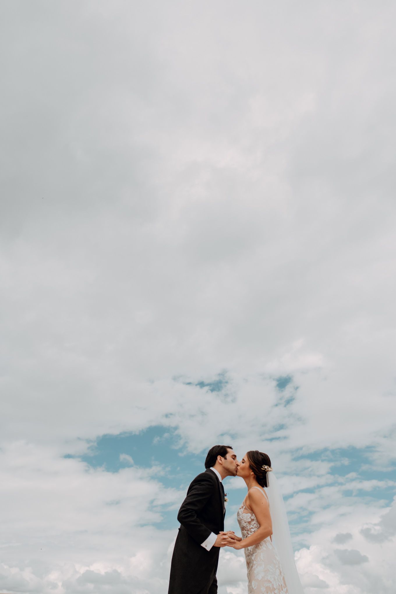 A couple embraces and kisses against a cloudy sky with patches of blue peeking through.