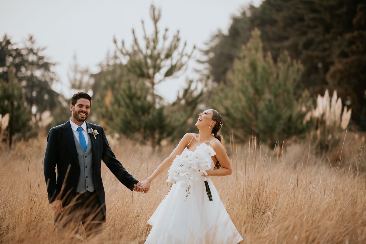 A happy couple walks hand in hand through a field of tall golden grass during their sunset wedding photo session.