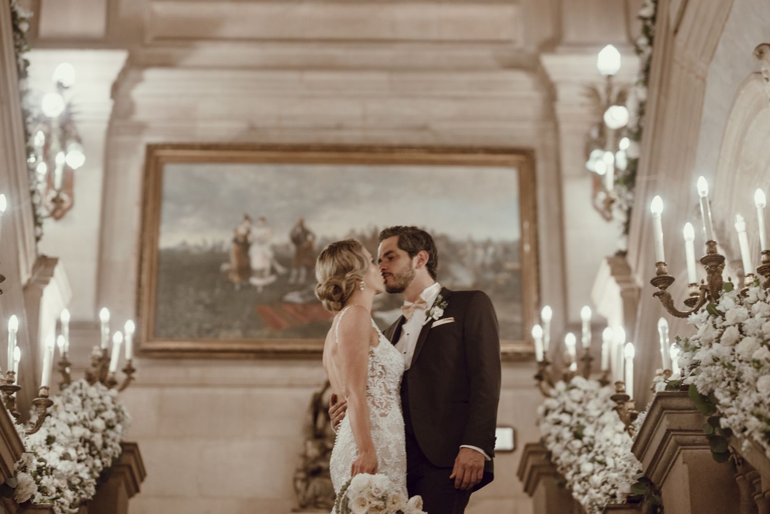 A romantic wedding moment captured in a grand hallway with ornate lighting and white floral decorations.