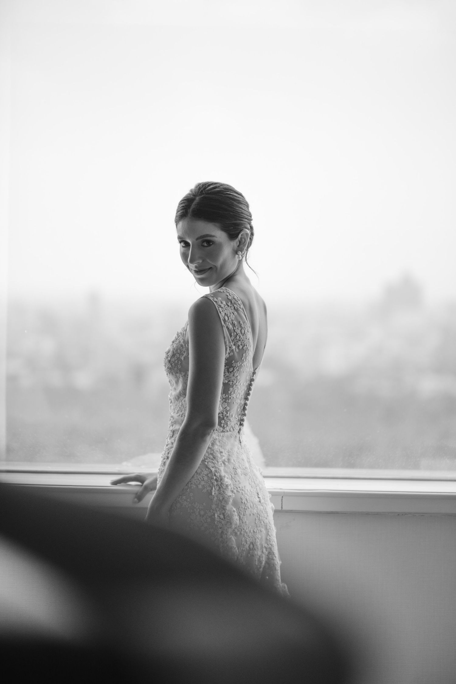 A classic black and white photo of a bride in a lace wedding dress posed against a window.