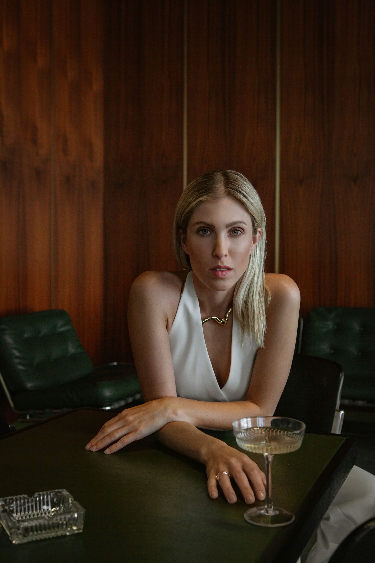 Woman in sleeveless white top with statement necklace seated at dark wood table with cocktail.