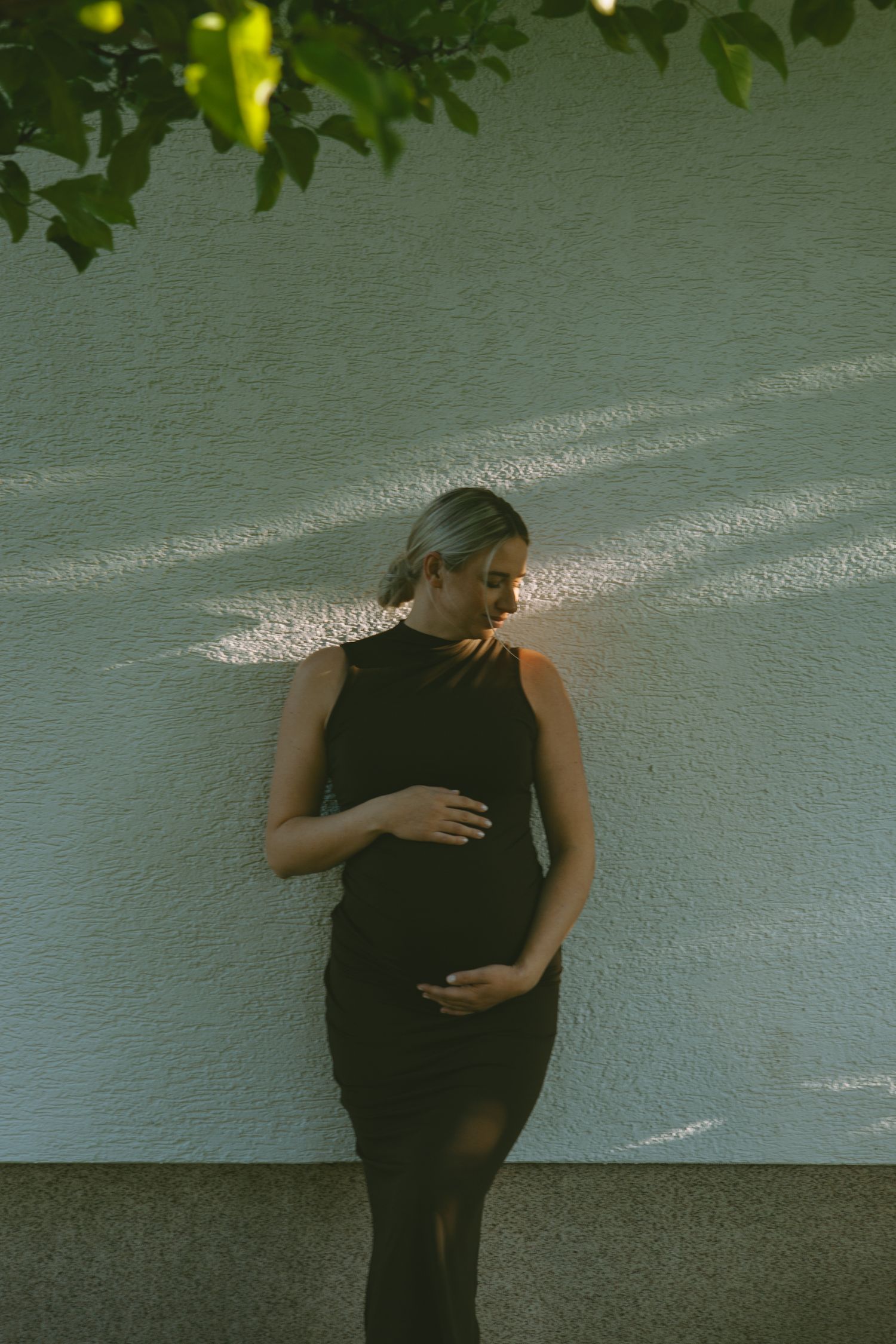 Figure in black clothing against white wall with leaves casting shadows from above.