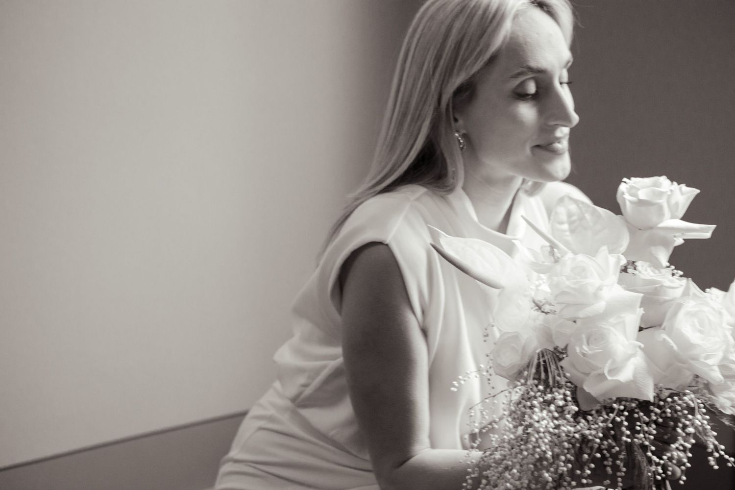 Elegant woman in white dress holding romantic bouquet near window in black and white photo.