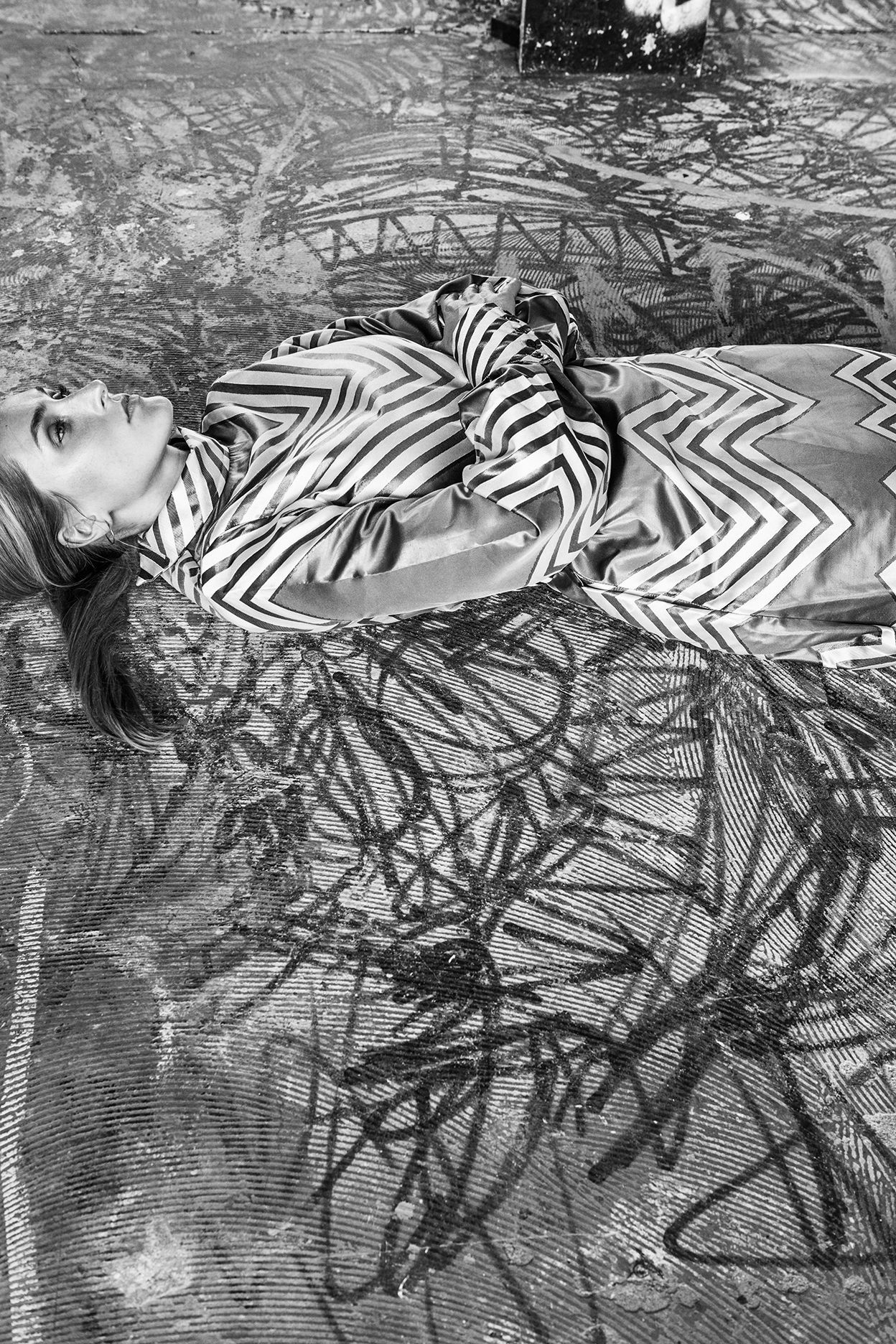 Black and white image showing a person lying down wearing a zigzag patterned dress against an abstract background.