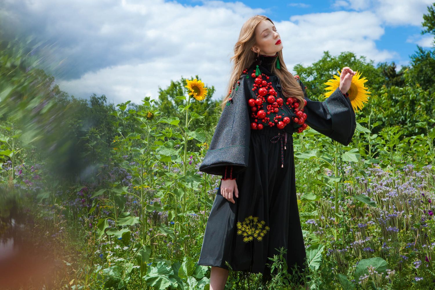 Whimsical garden fashion shoot with black coat and red floral accents among blooming sunflowers.
