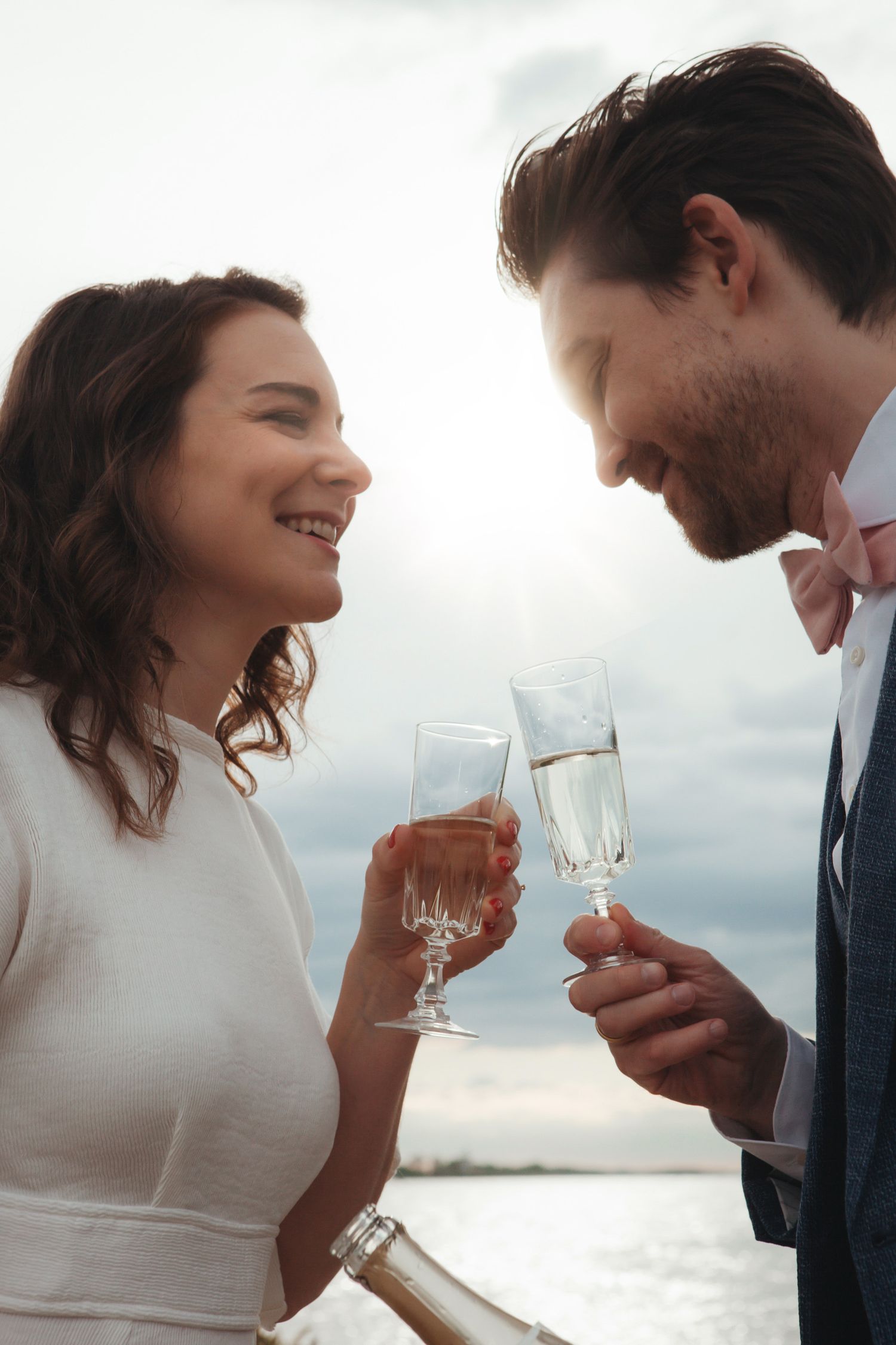 A couple smiles while toasting champagne glasses at sunset by the water.