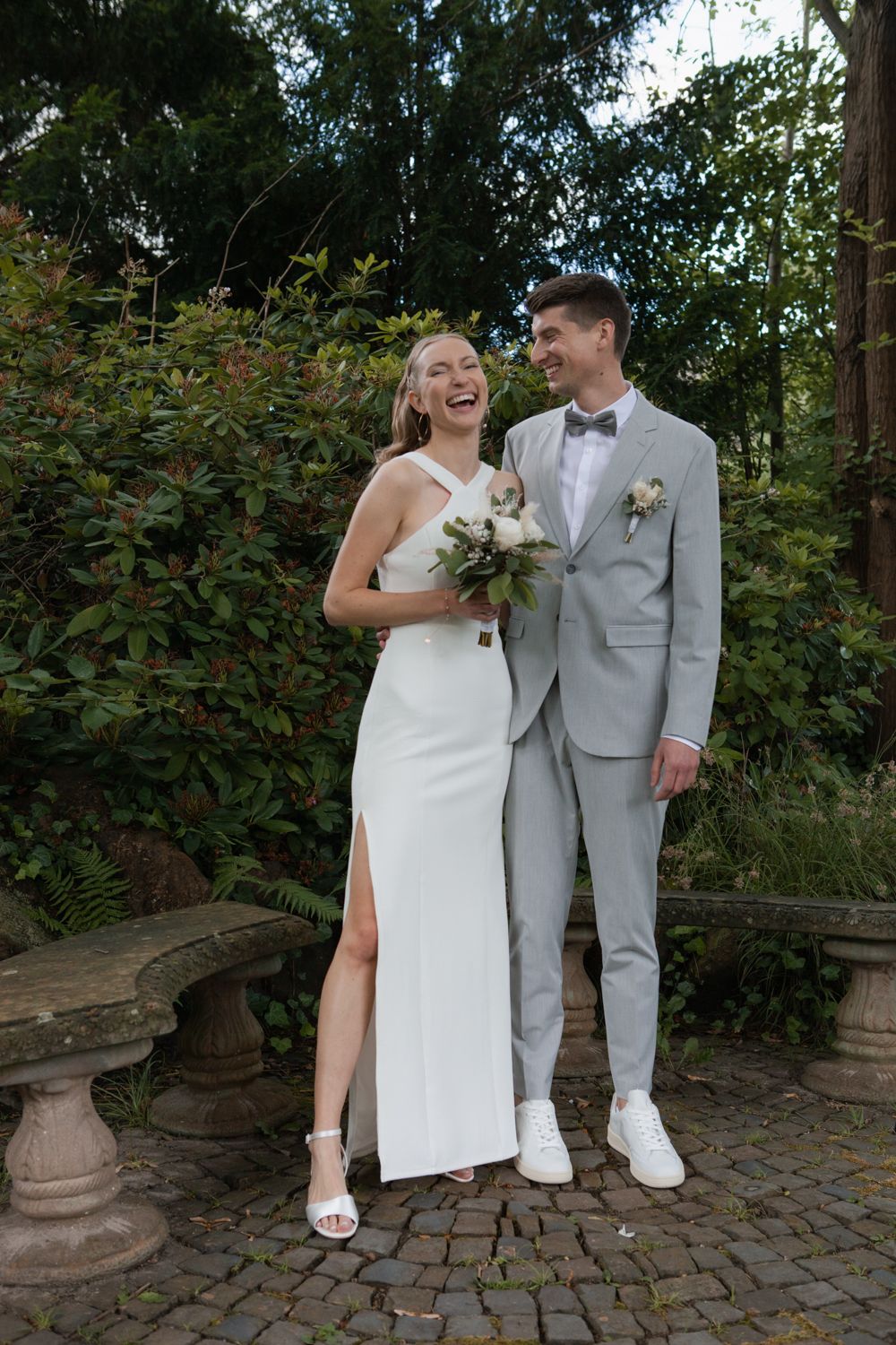 A couple in wedding attire stands on cobblestone, wearing matching white outfits and sharing a joyful moment outdoors.
