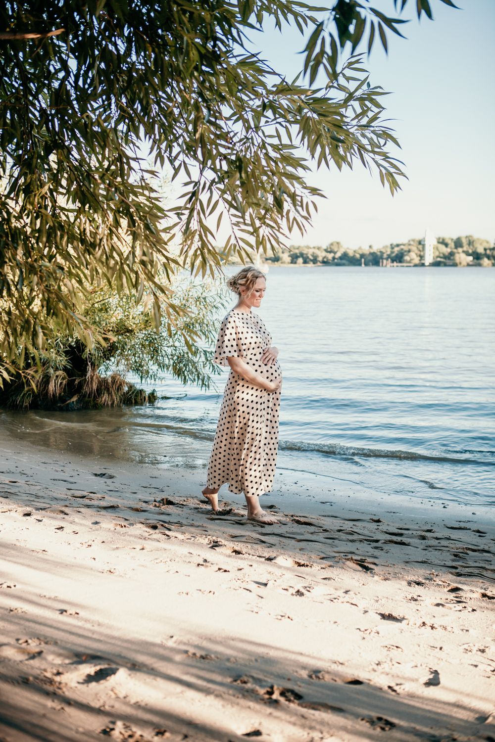 A woman in a polka dot dress stands by the waterfront under hanging willow branches.