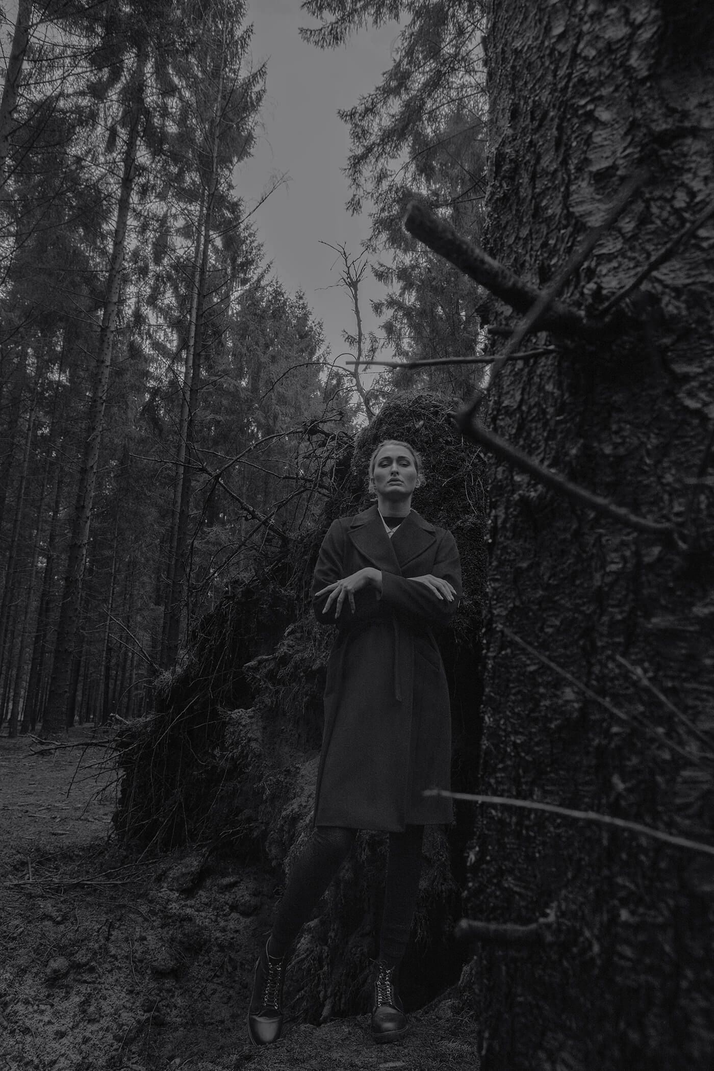 A solitary figure in a dark coat poses against a tree in a moody forest setting.