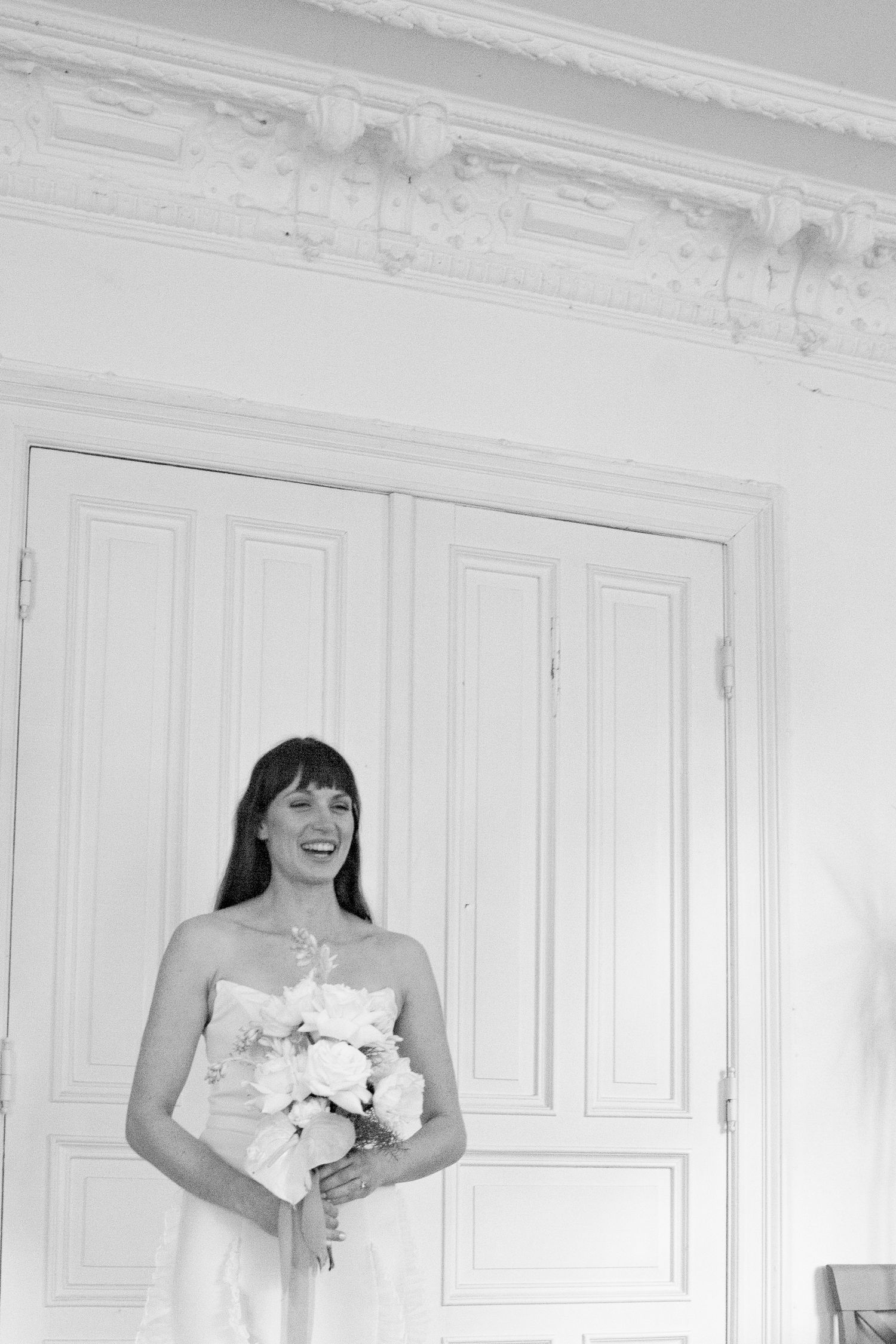 A joyful bride in white holds a bouquet while standing against an ornate white wall.