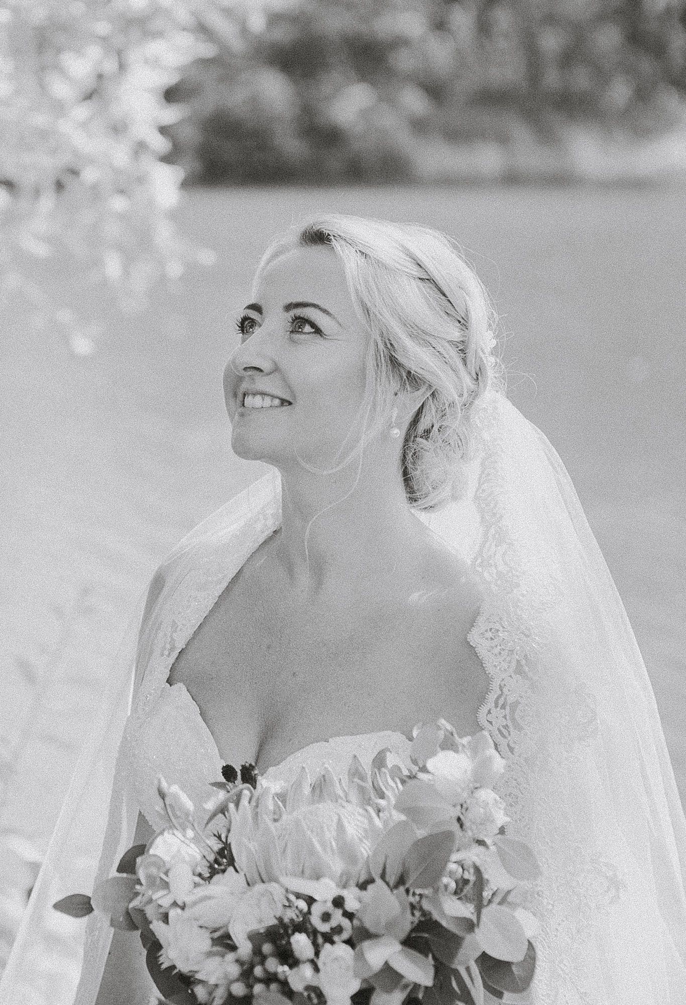 Black and white photo of a joyful bride wearing a strapless gown and veil while holding a floral bouquet outdoors.