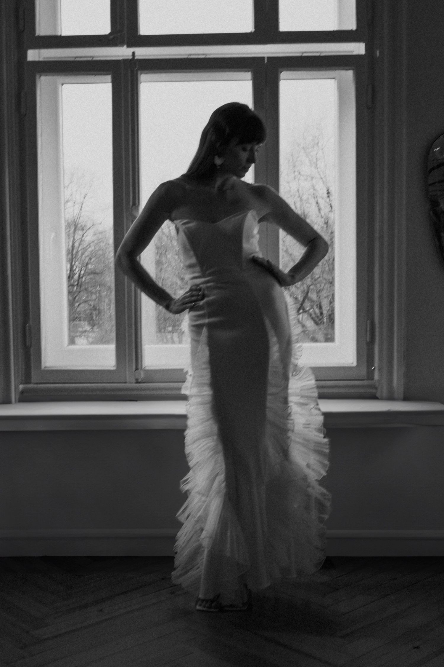 Silhouetted figure in a fitted wedding gown poses dramatically by a window.