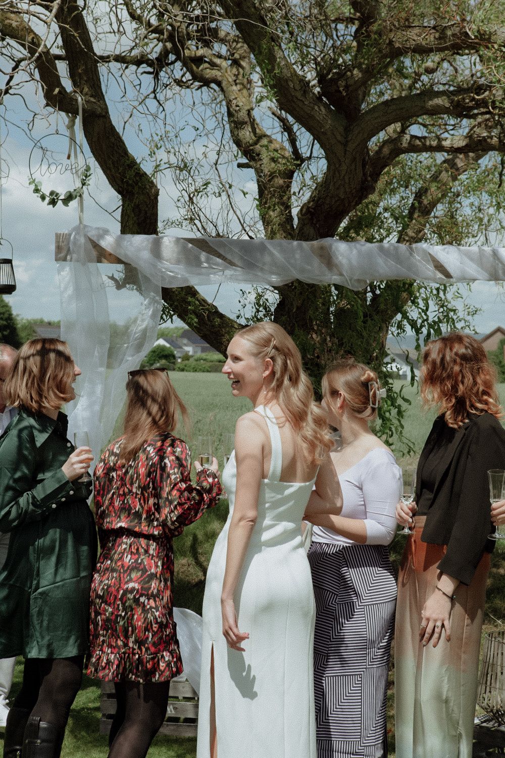 A group of people in formal attire gathers under a tree for a photo shoot during an outdoor celebration.