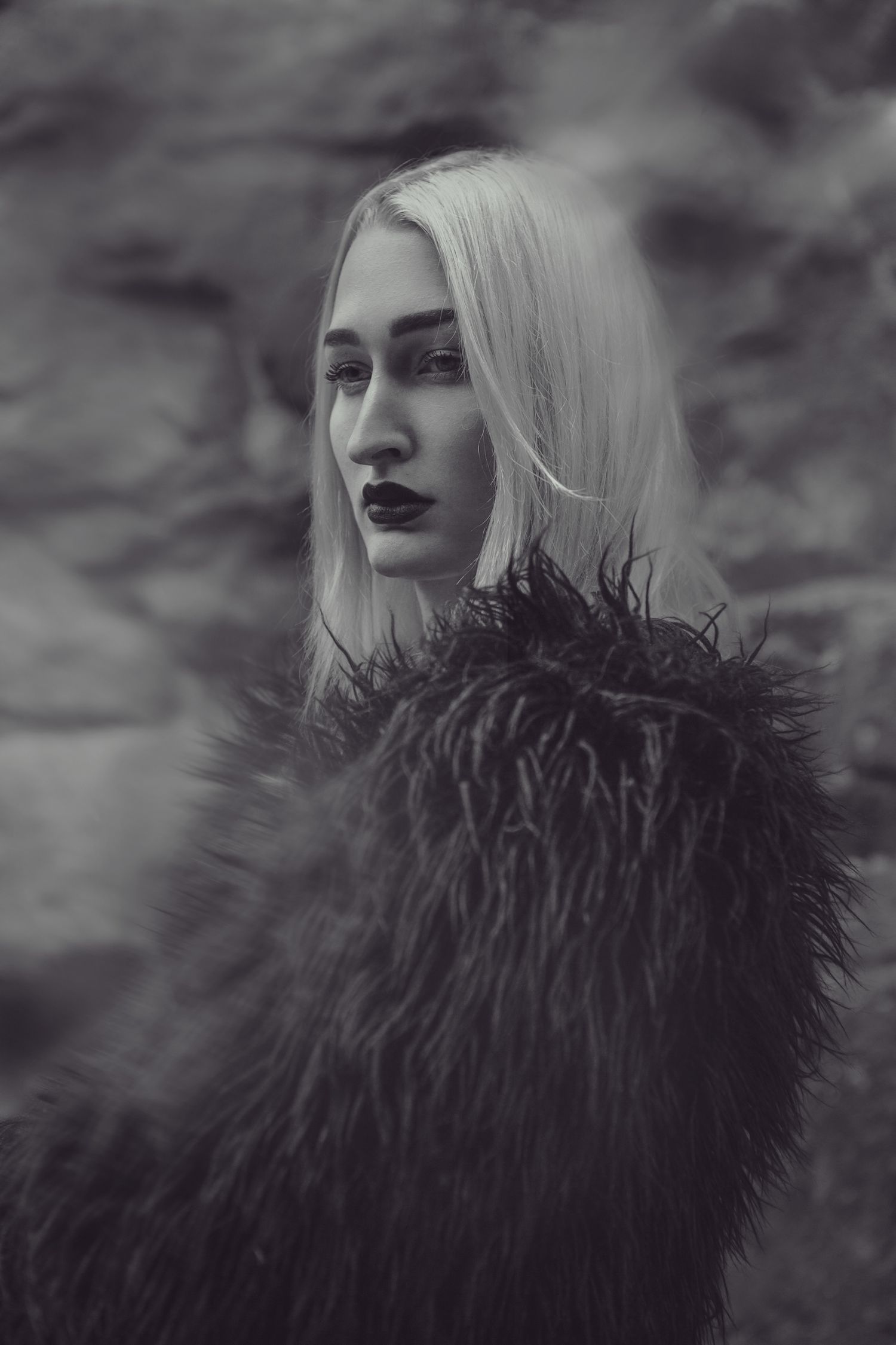 Moody portrait featuring a person wearing a black furry coat against cloudy background.
