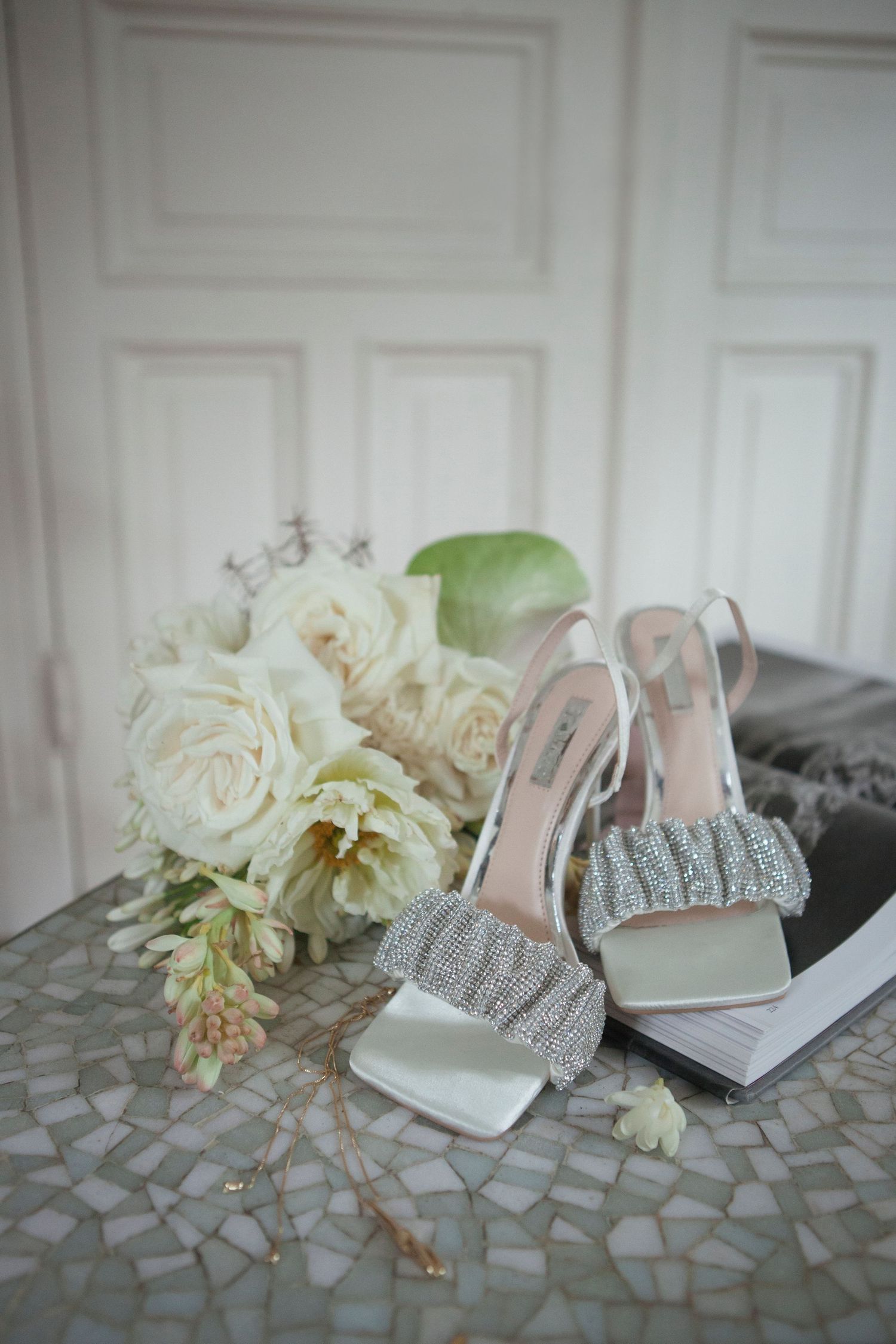 Elegant white bridal shoes with crystal embellishments alongside a bouquet of cream roses on a mosaic table surface.