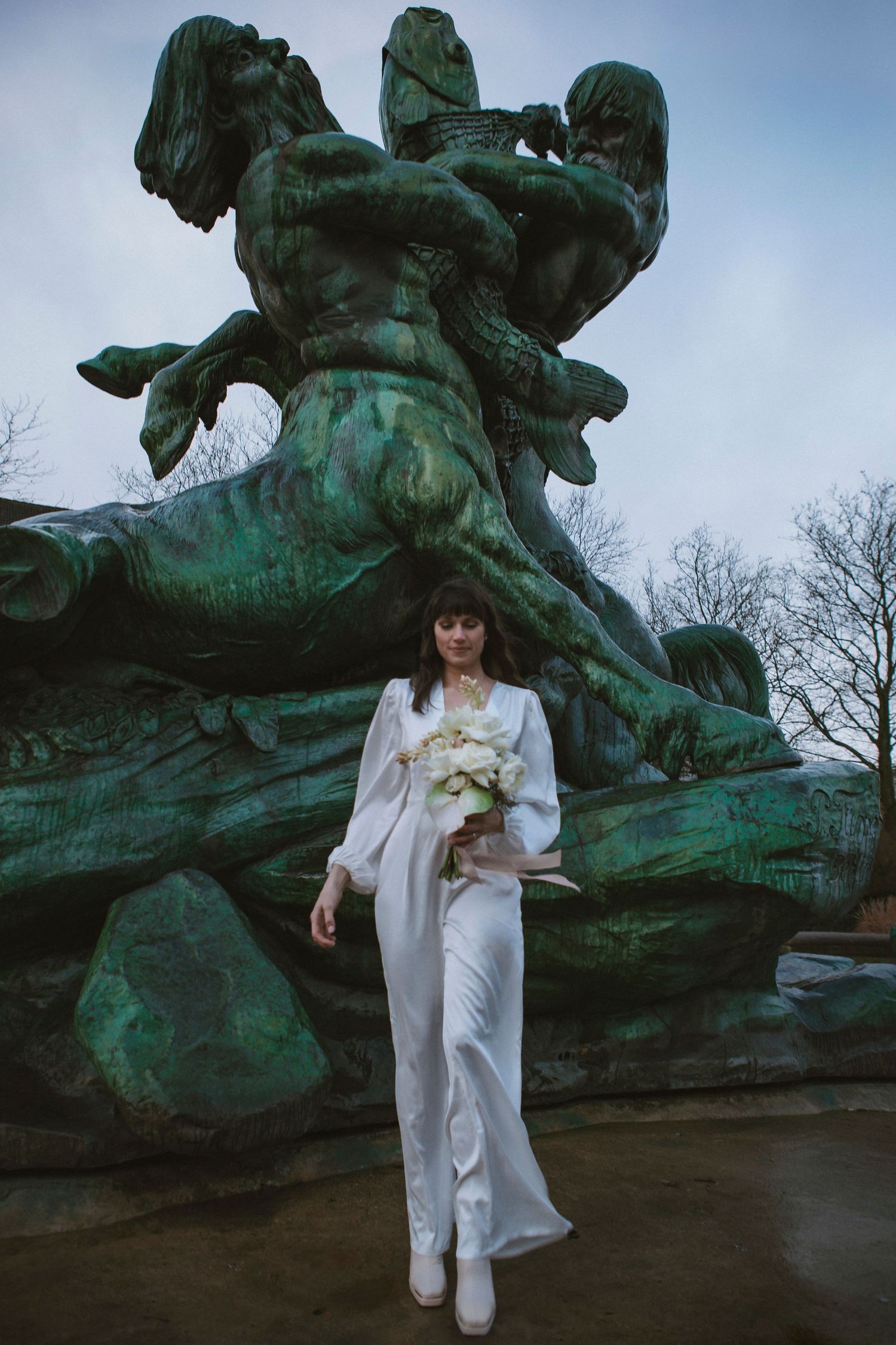 Person in white outfit sits near large green bronze sculpture in outdoor setting at dusk.
