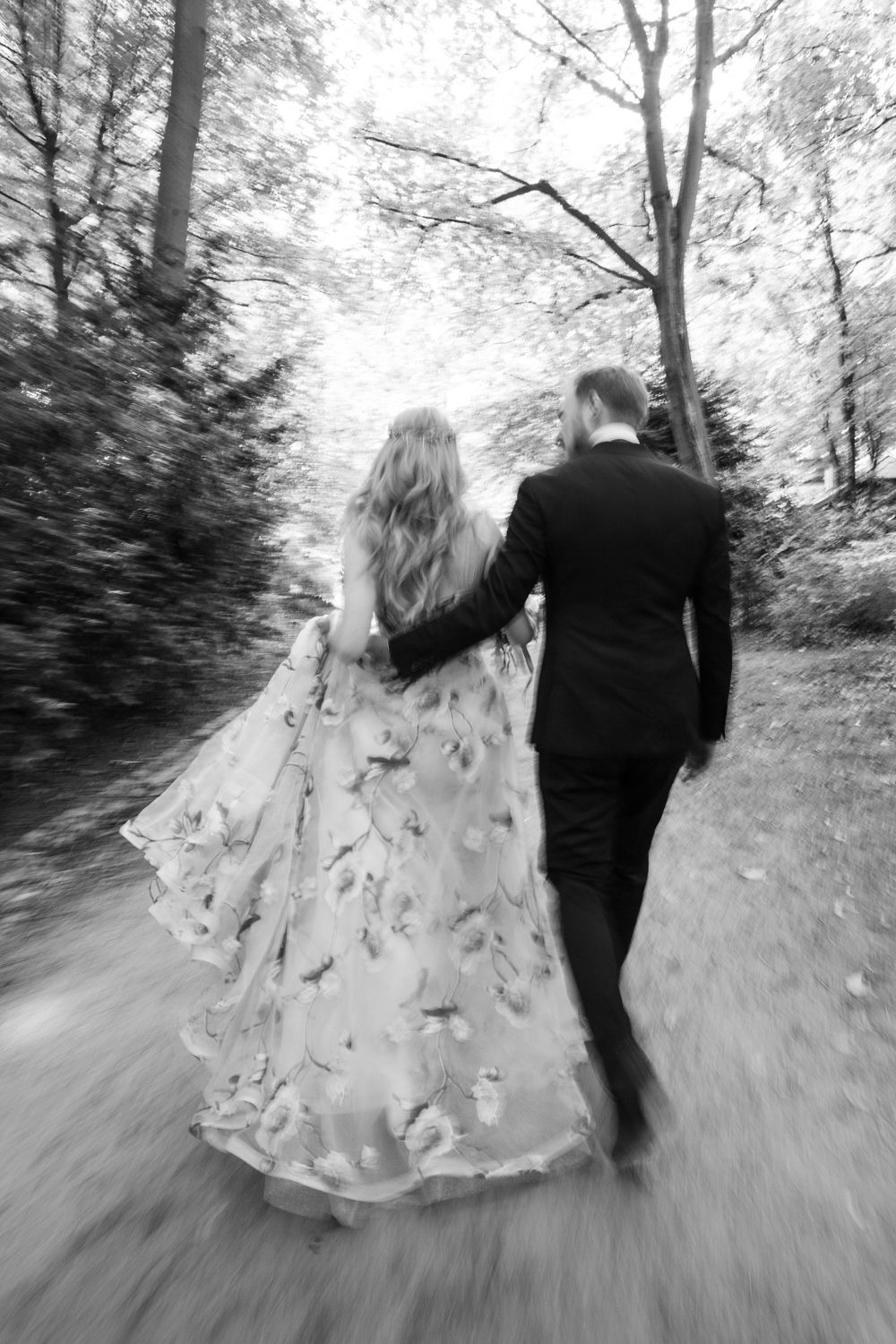 A couple in formal attire walking down a wooded path, with the bride's flowing floral dress trailing behind in black and white.