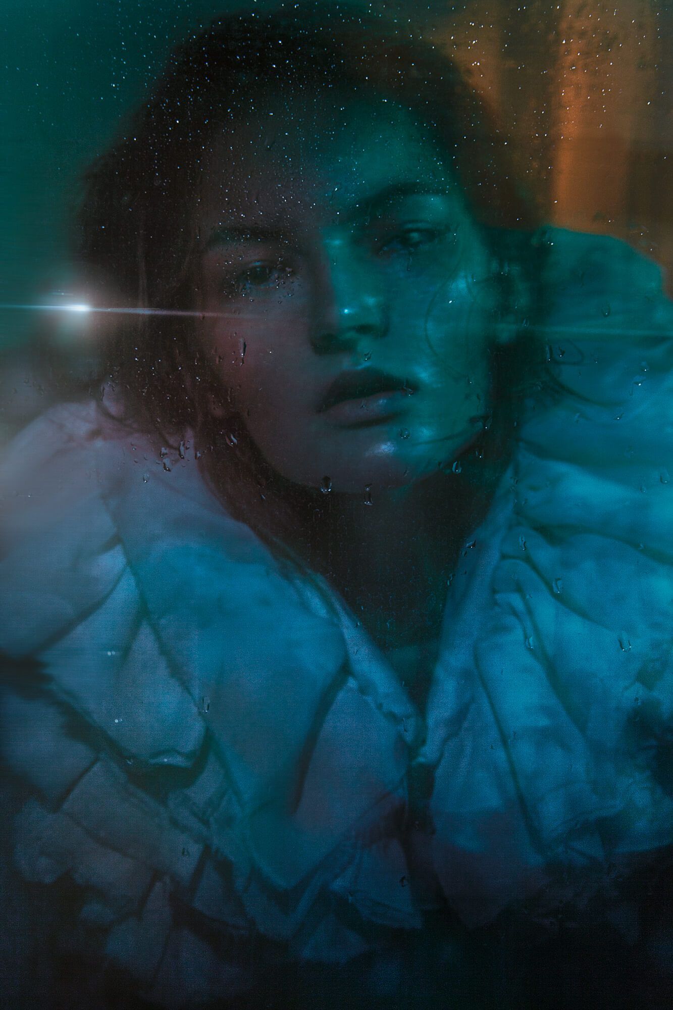 Moody portrait with teal and orange lighting through wet glass.