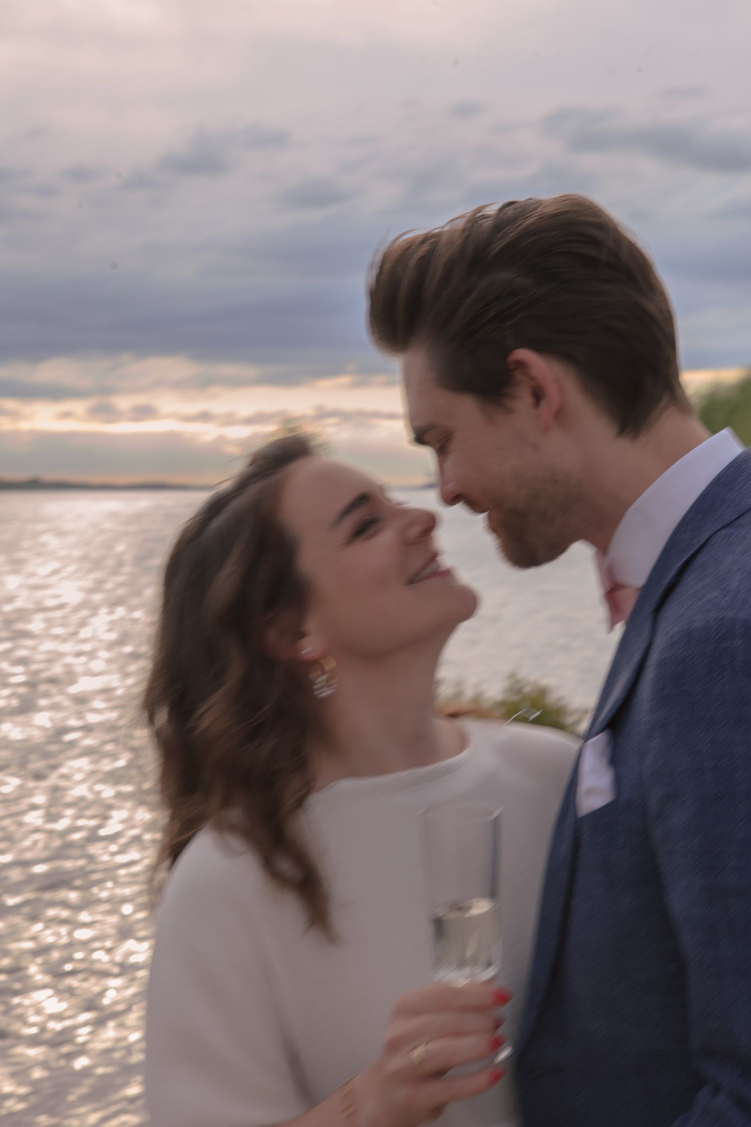 A romantic couple shares an intimate moment at sunset by the water's edge while holding champagne glasses.