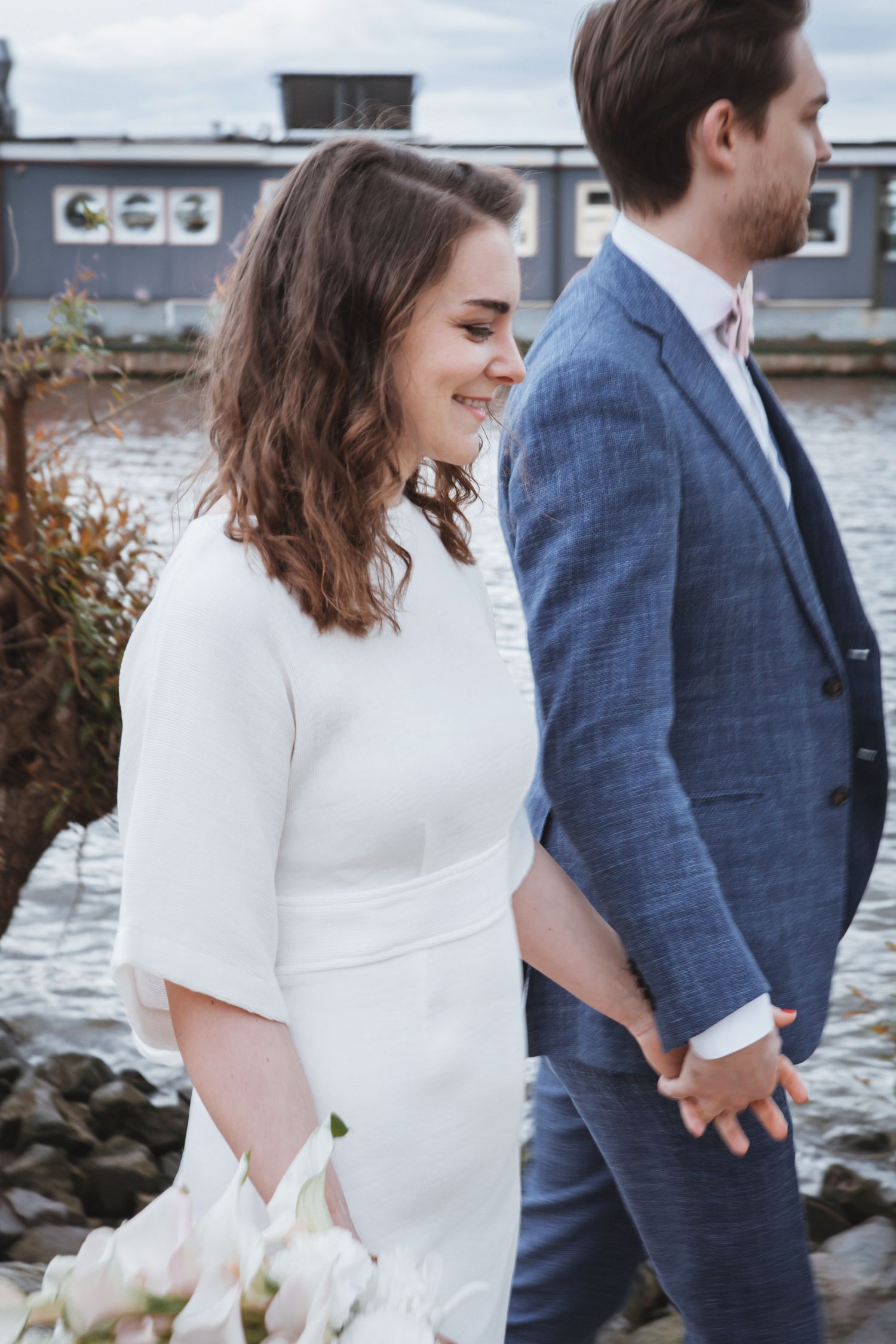 A couple in wedding attire stand by the water, with the bride in white and groom in a blue suit holding hands.