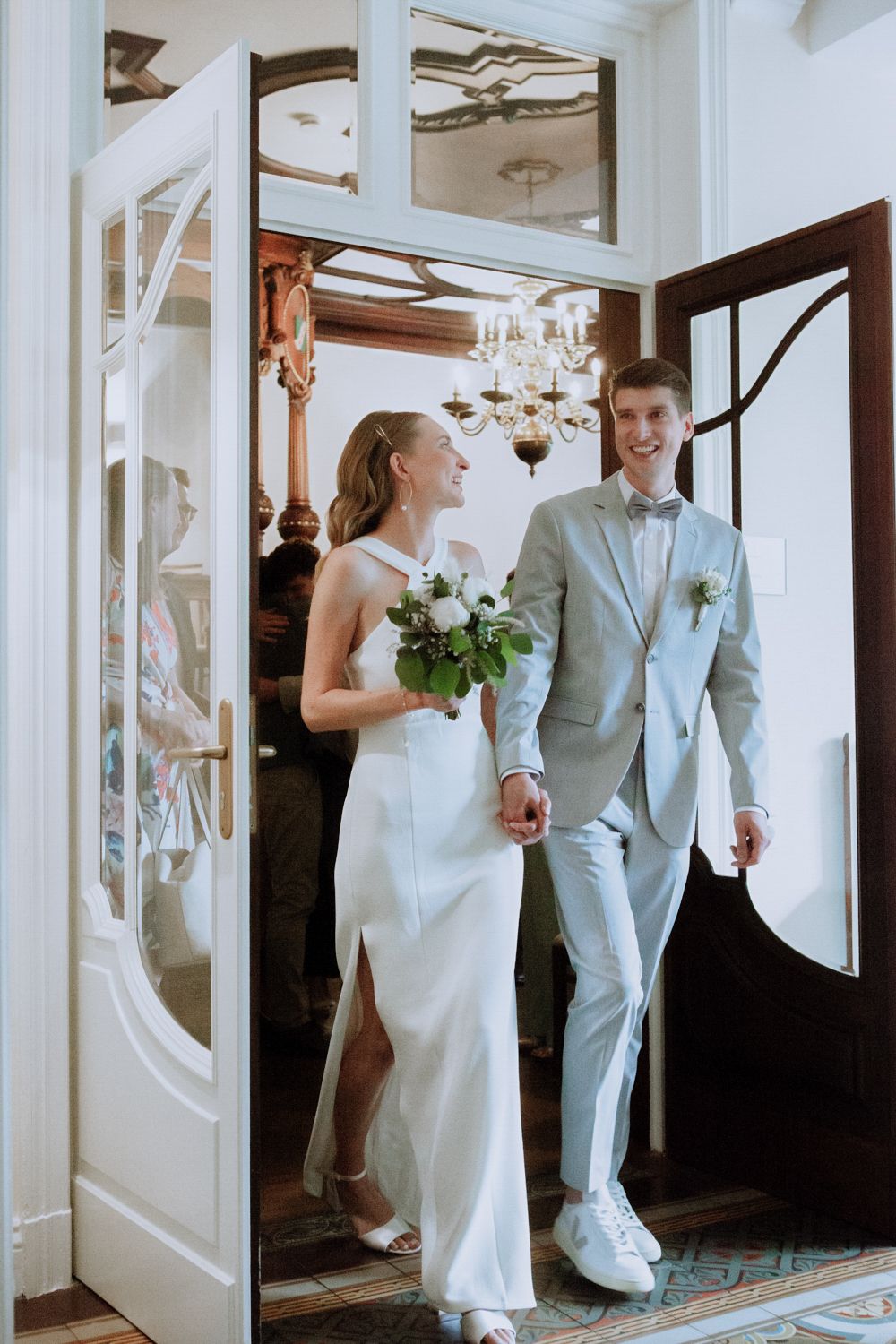 A couple in wedding attire walk through an elegant doorway with white paneling and a crystal chandelier.