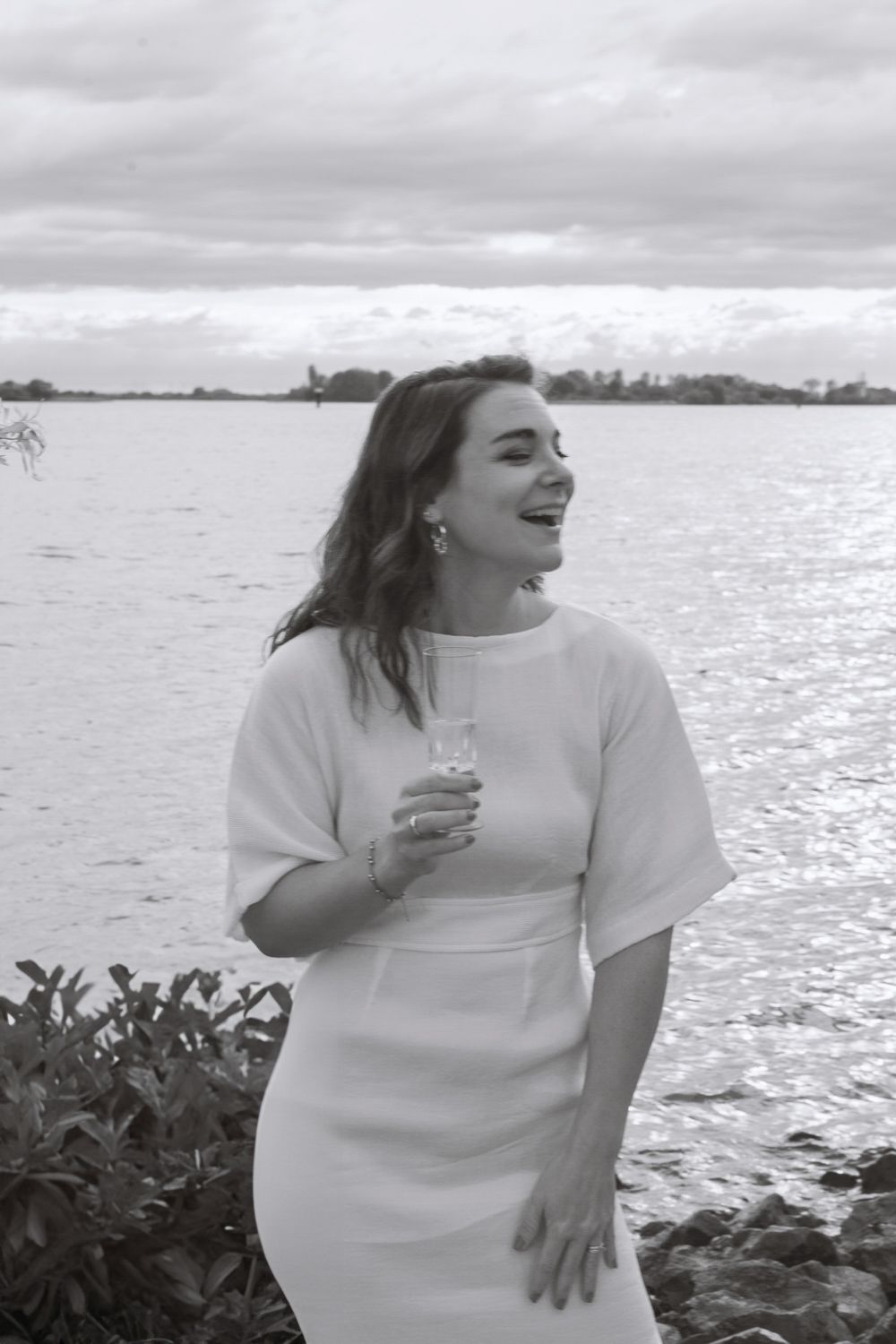 A black and white photograph showing a figure in a white dress standing by a waterfront on an overcast day.