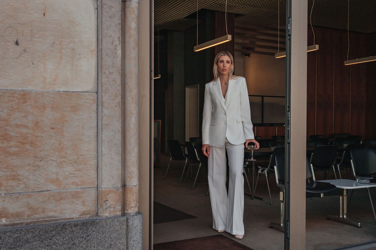Fashionable figure in white pantsuit standing in doorway of modern building.