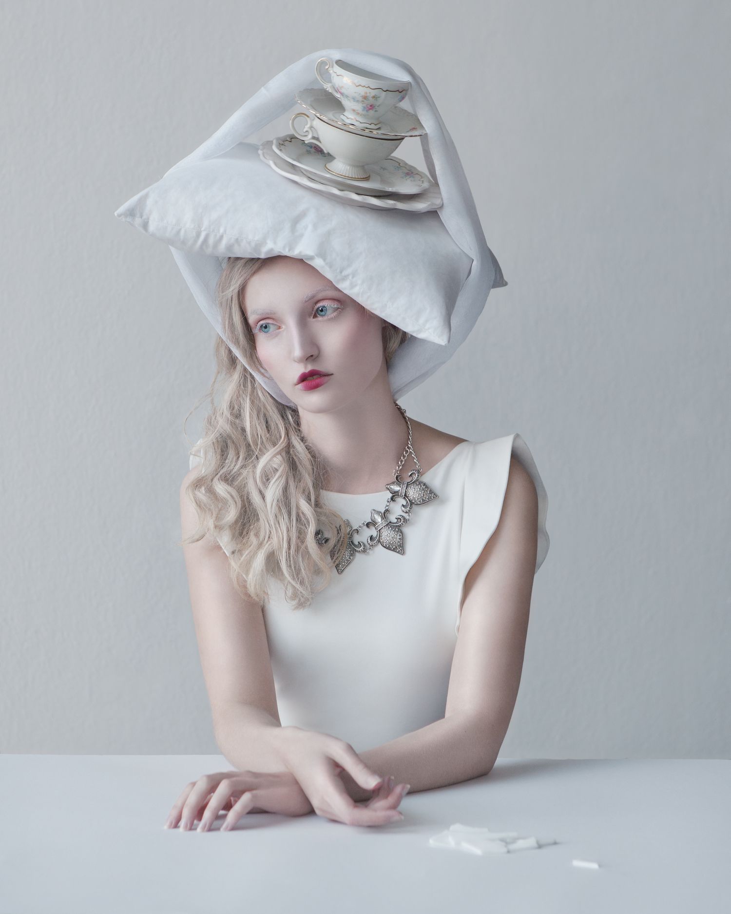 Surreal portrait featuring a person wearing teacup hat and white dress against light background.