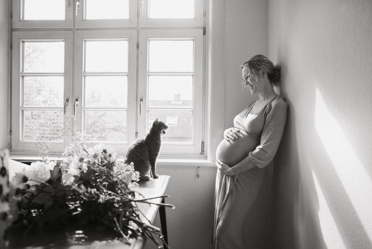 Black and white photo of a pregnant woman standing by a bright window with a black cat perched on a floral surface.