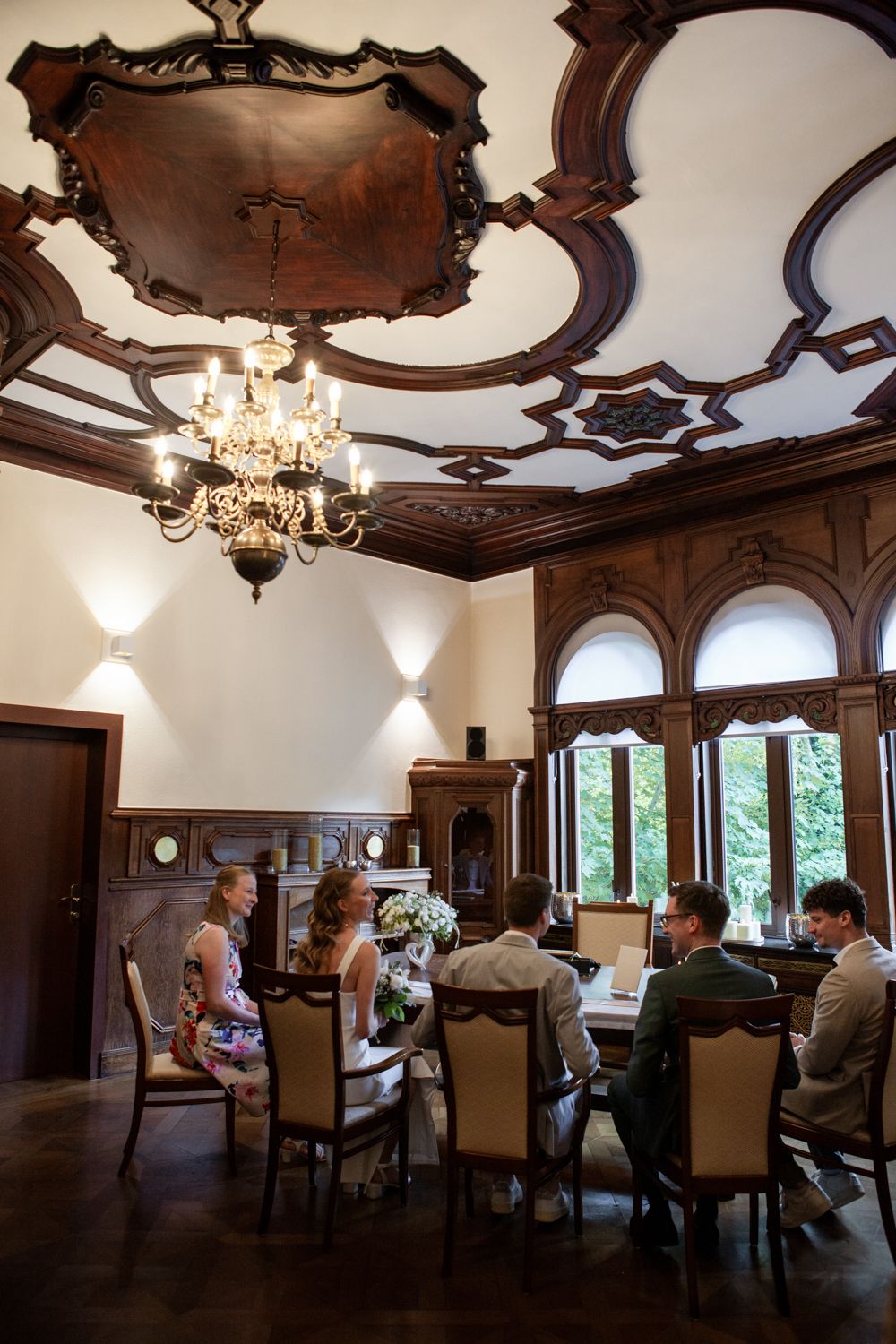 Elegant dining room with ornate wood ceiling, crystal chandelier and arched windows in a historic mansion.