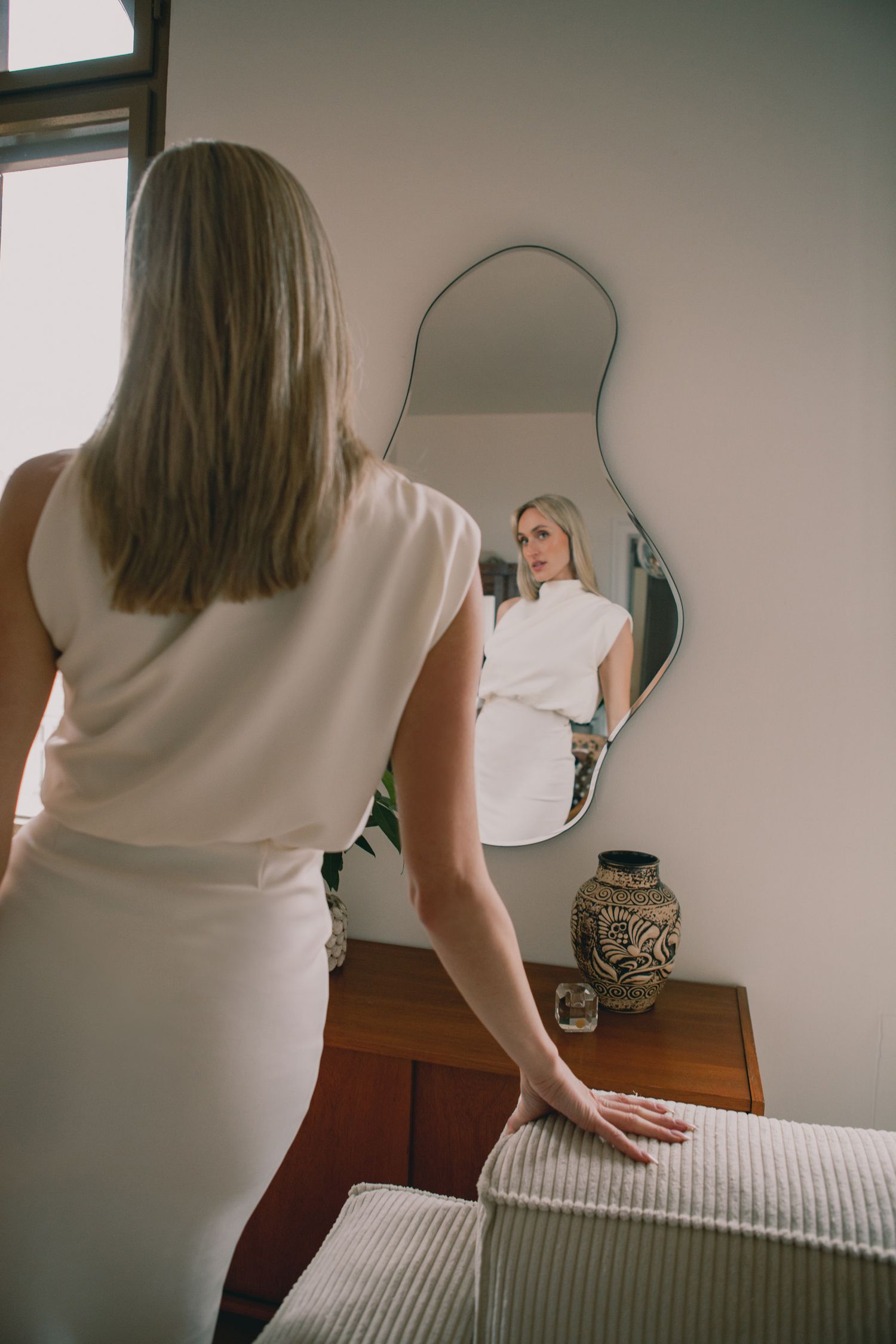 Bride in sleek white dress looking at reflection in curved wall mirror.