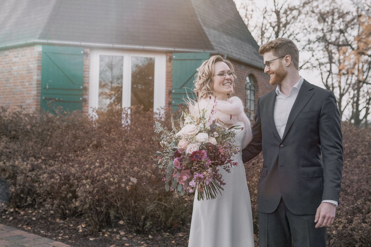 Couple sharing a romantic moment in an autumn garden setting with rustic farmhouse in the background.
