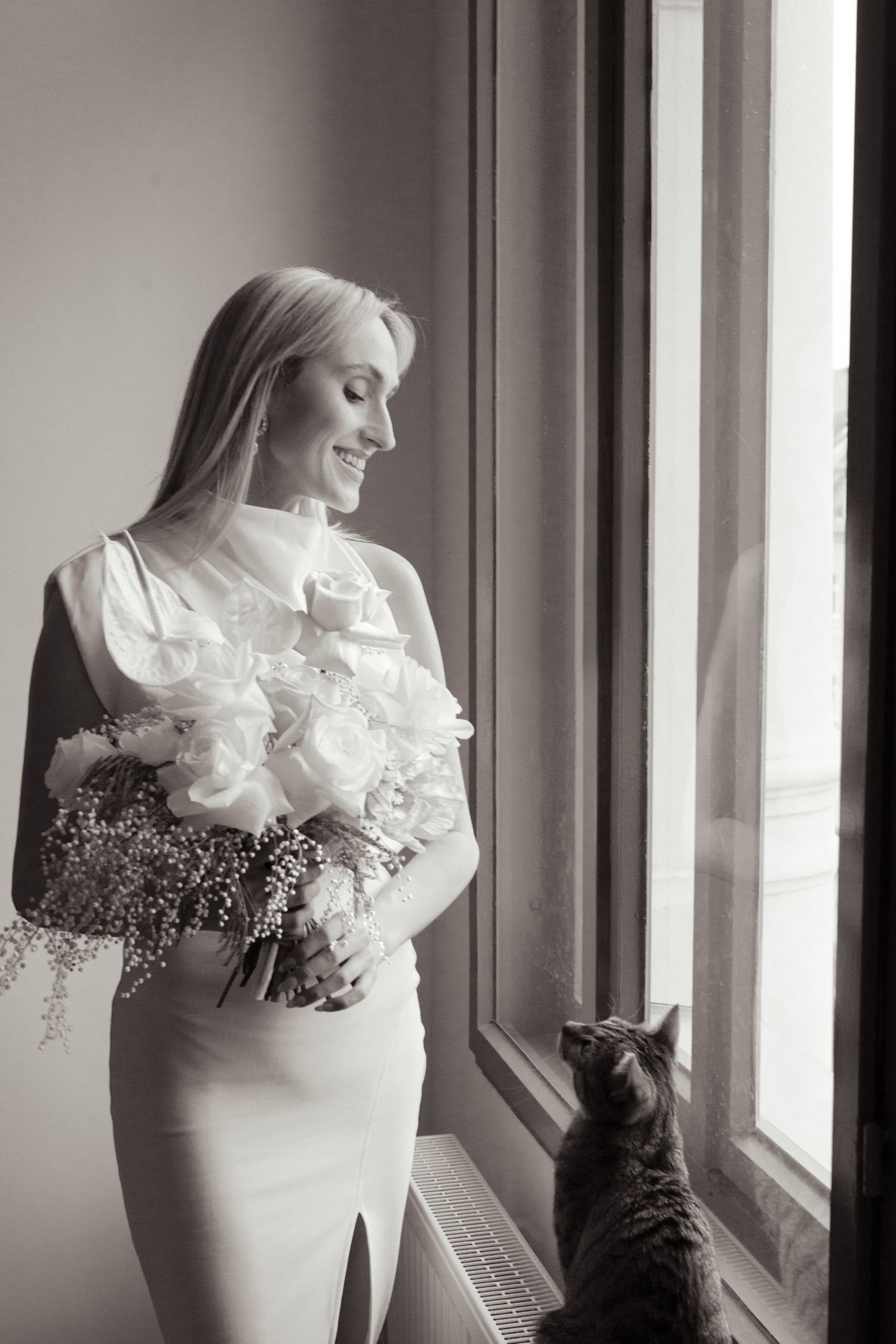 Black and white photo showing a bride with flowers looking out window while cat watches nearby.