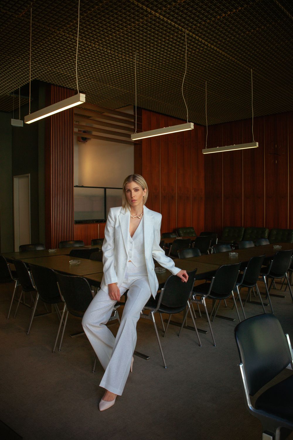 A person in a crisp white suit sits casually on chairs in a dimly lit auditorium setting.
