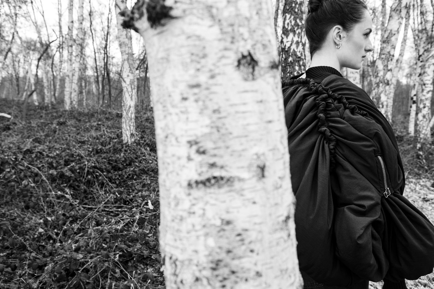 Black and white photo showing a figure standing by a birch tree trunk in a forest.