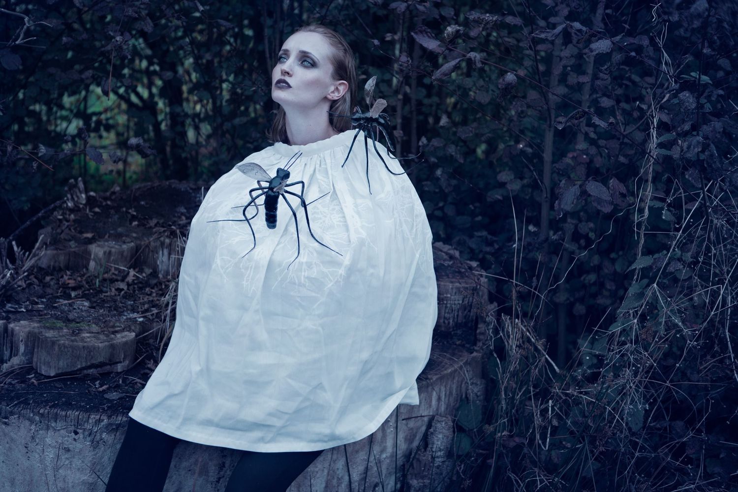 Figure in white wearing a spider-decorated cape stands against a dark forest backdrop.