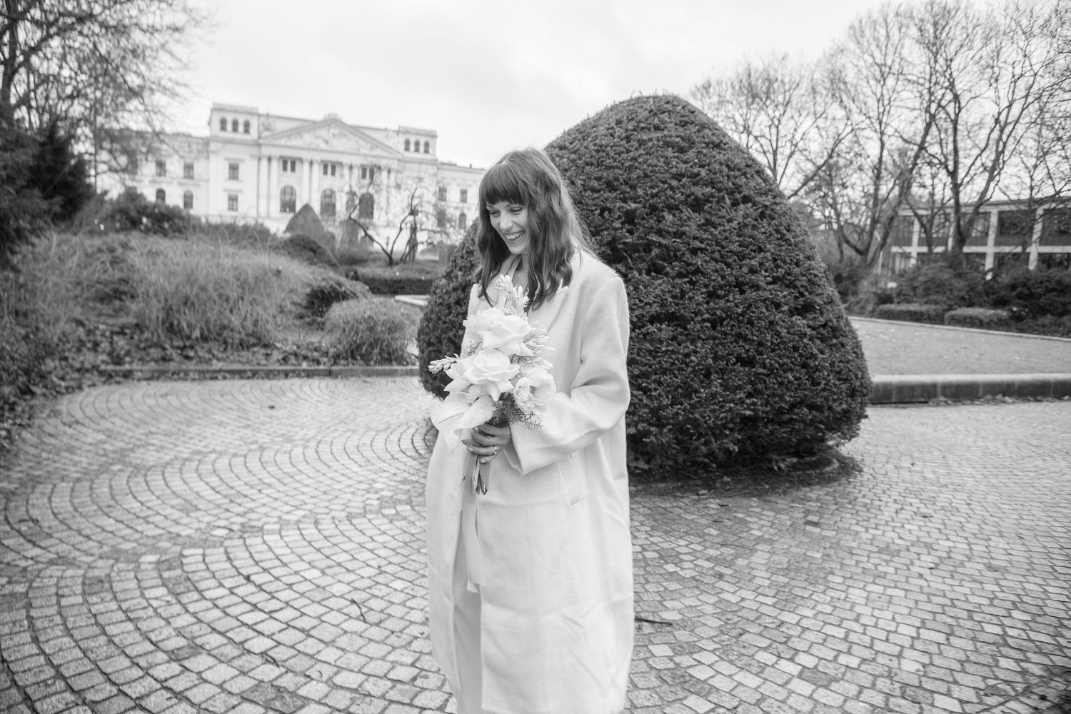 Black and white portrait of person in white coat holding flowers in garden setting.