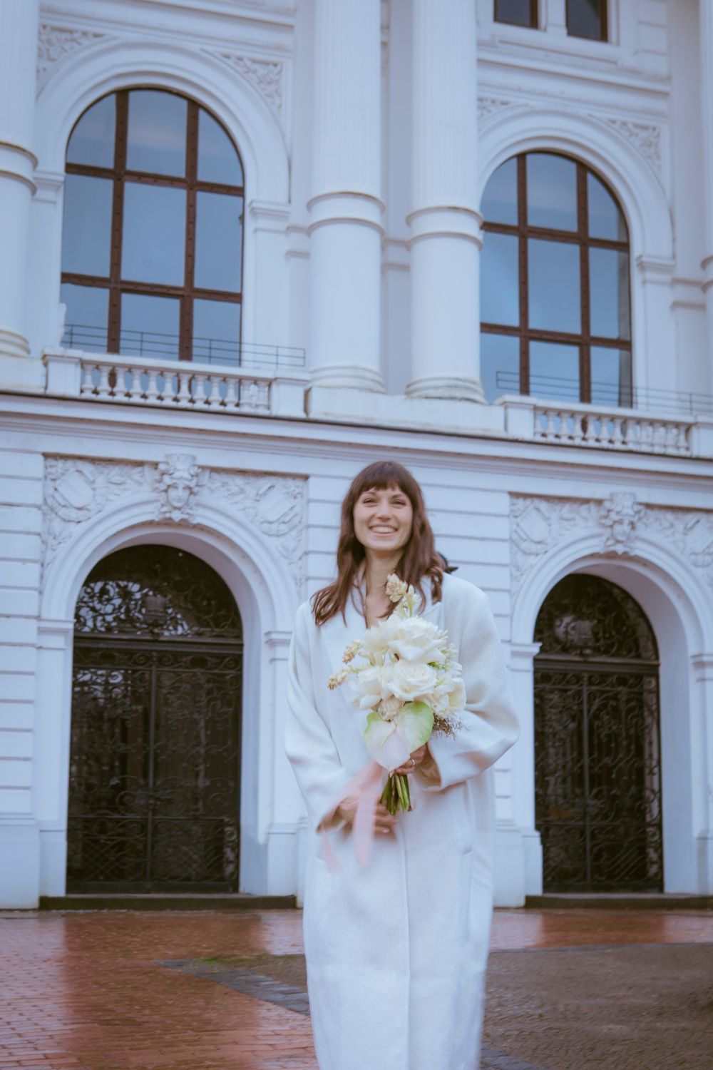 A bride in a white dress stands on a terrace with arched windows and classical architecture in the background.