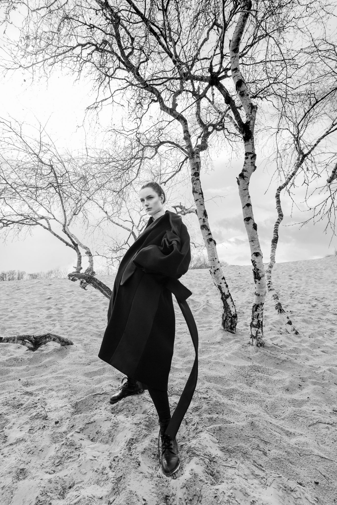 Fashion figure in long black coat poses among bare birch trees in winter landscape.