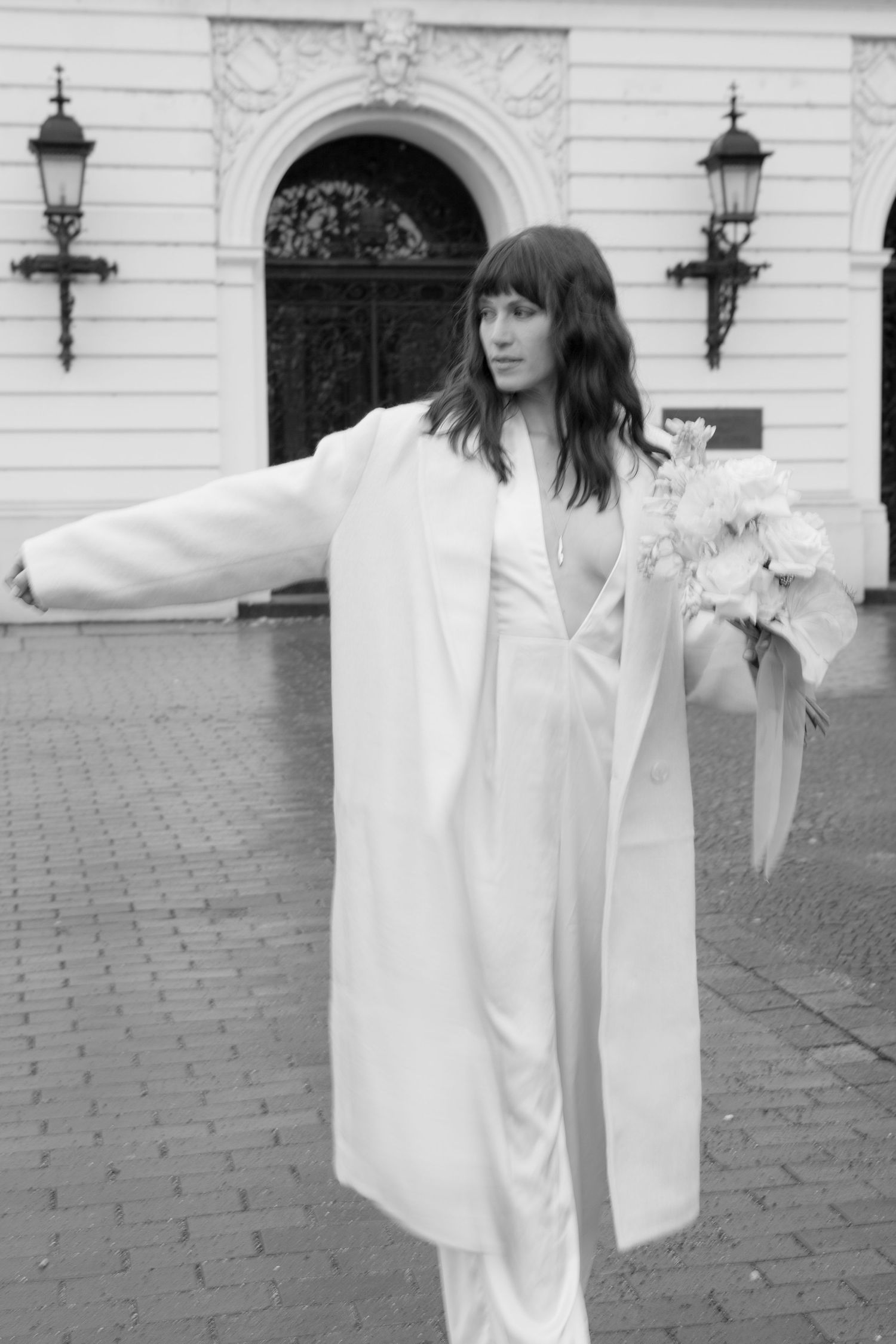Monochrome photo of bride in white pantsuit and long coat holding flowers against historic building.