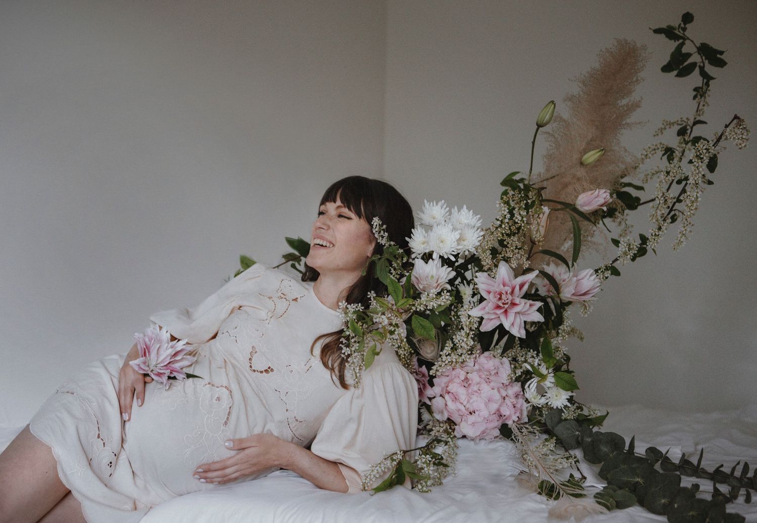 Dreamy maternity scene with flowers and woman in white dress reclining among pink blooms.